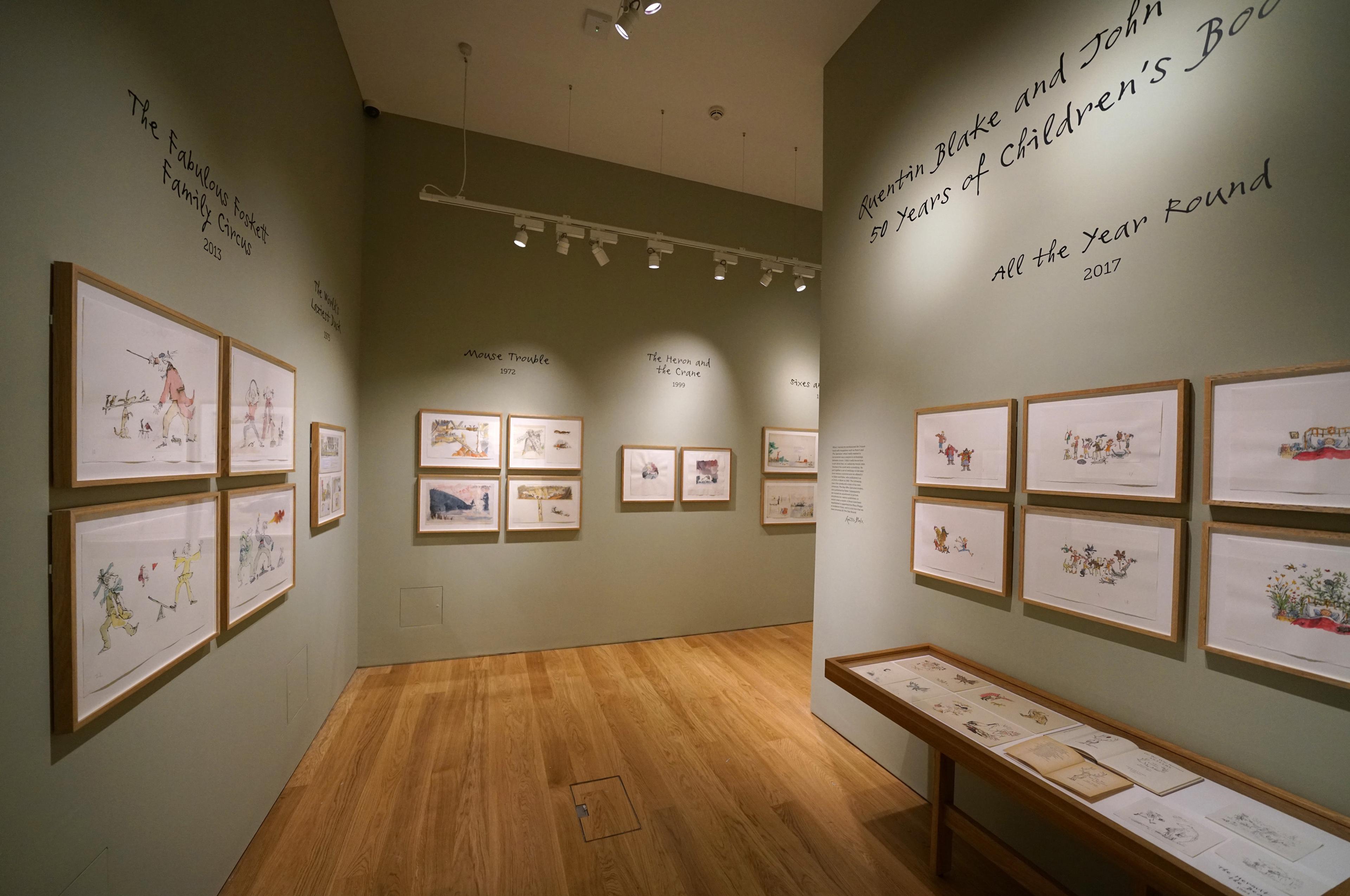 Framed illustrations and text displayed on a green wall in a gallery space.