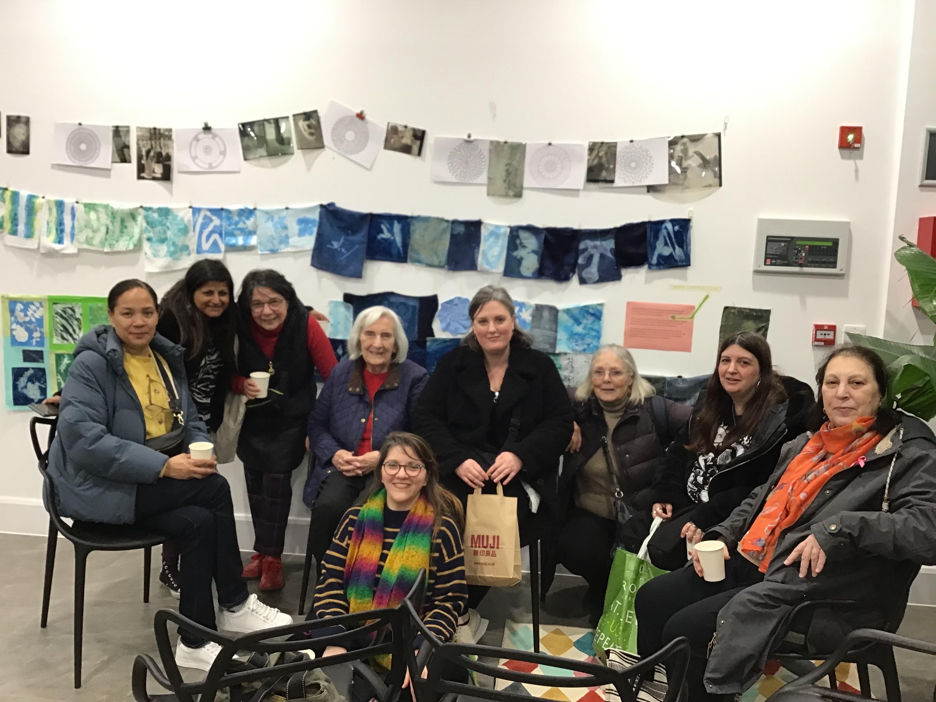 A photograph of nine people sitting in an exhibition space with fabric bunting hung up on the wall behind them.