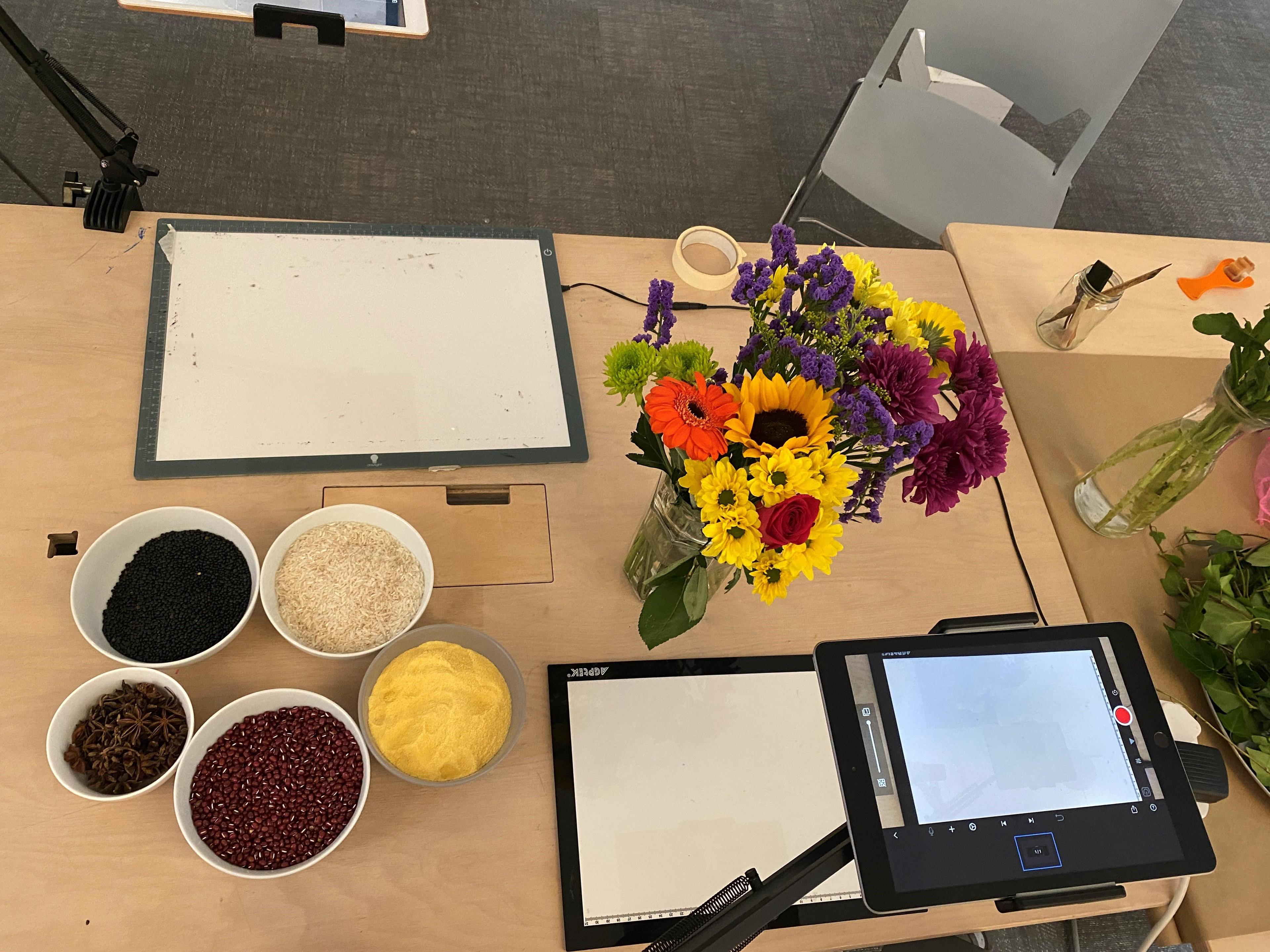 A photograph of the top of a desk with a vase of flowers on it, a digital tablet and bowls of sand and grains.