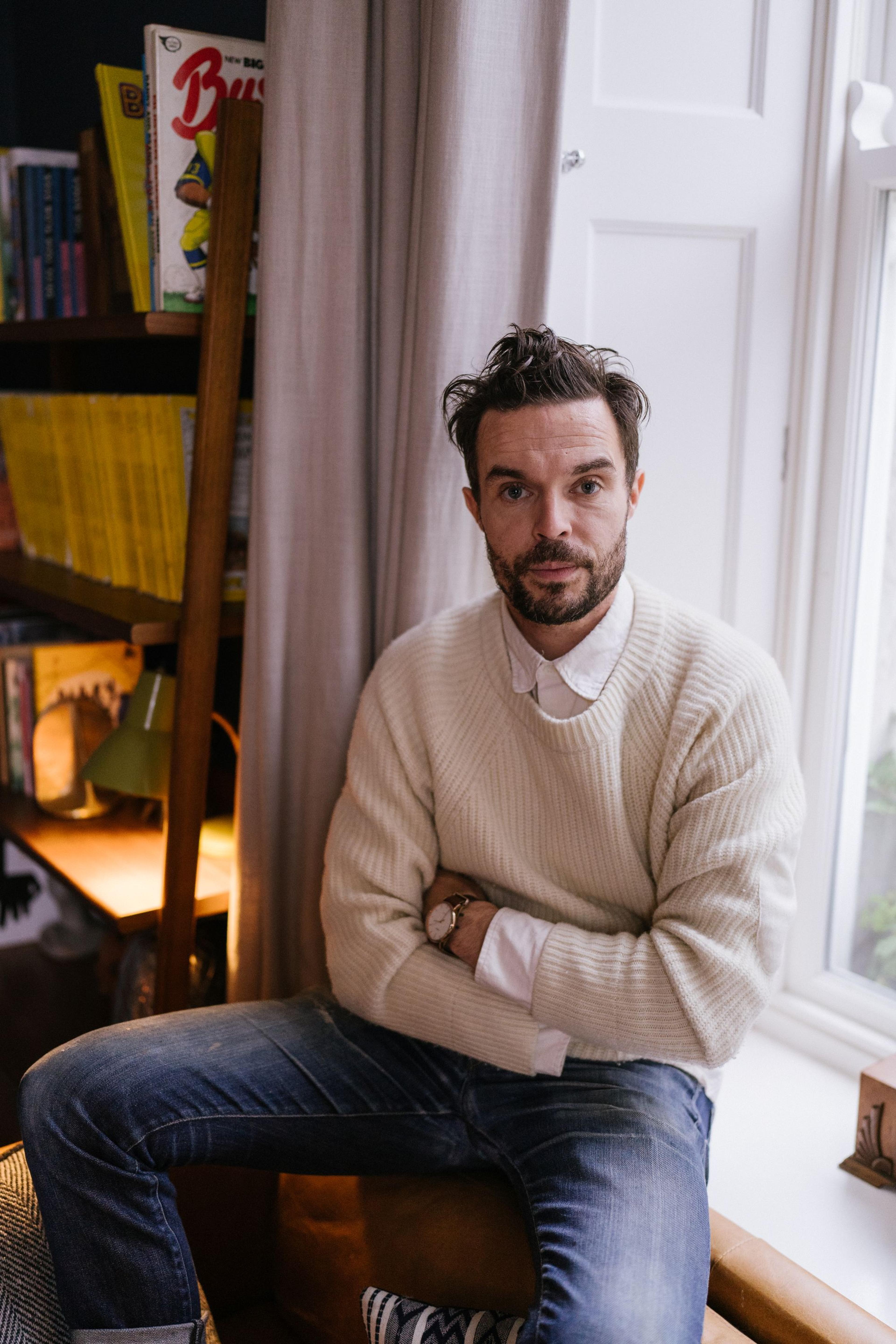 A photo of a man sitting on a window sill with crossed arms 