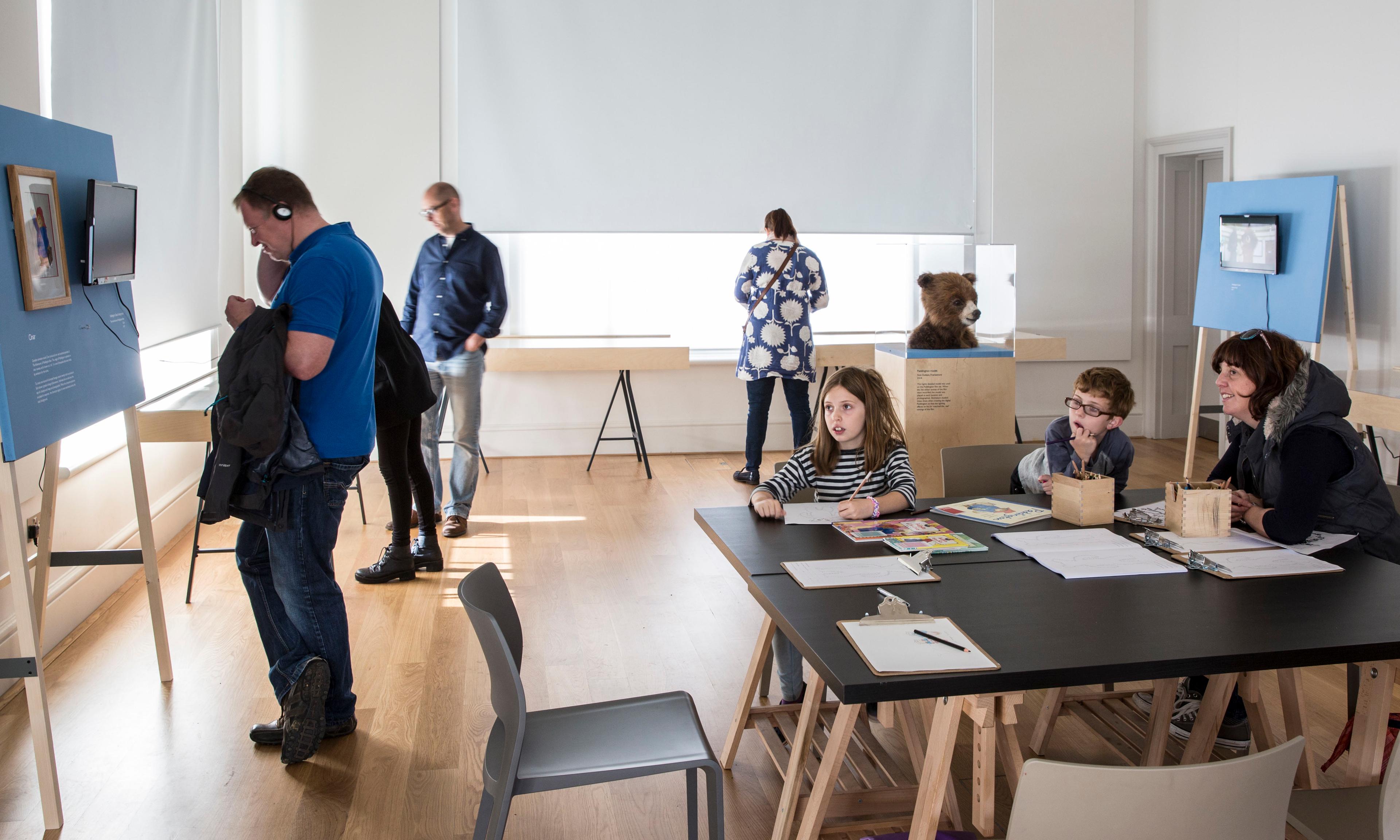 Adults and children in an exhibition space with tables, chairs and displays including a furry bear head is in a glass display case. 