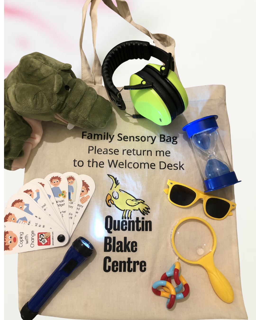 Tote bag reading "Family Sensory Bag, Please return to the Welcome Desk, Quentin Blake Centre" beneath a stuffed crocodile toy, emotion cards, a torch, a fidget toy, a magnifying glass, sunglasses, a sand timer and headphones.