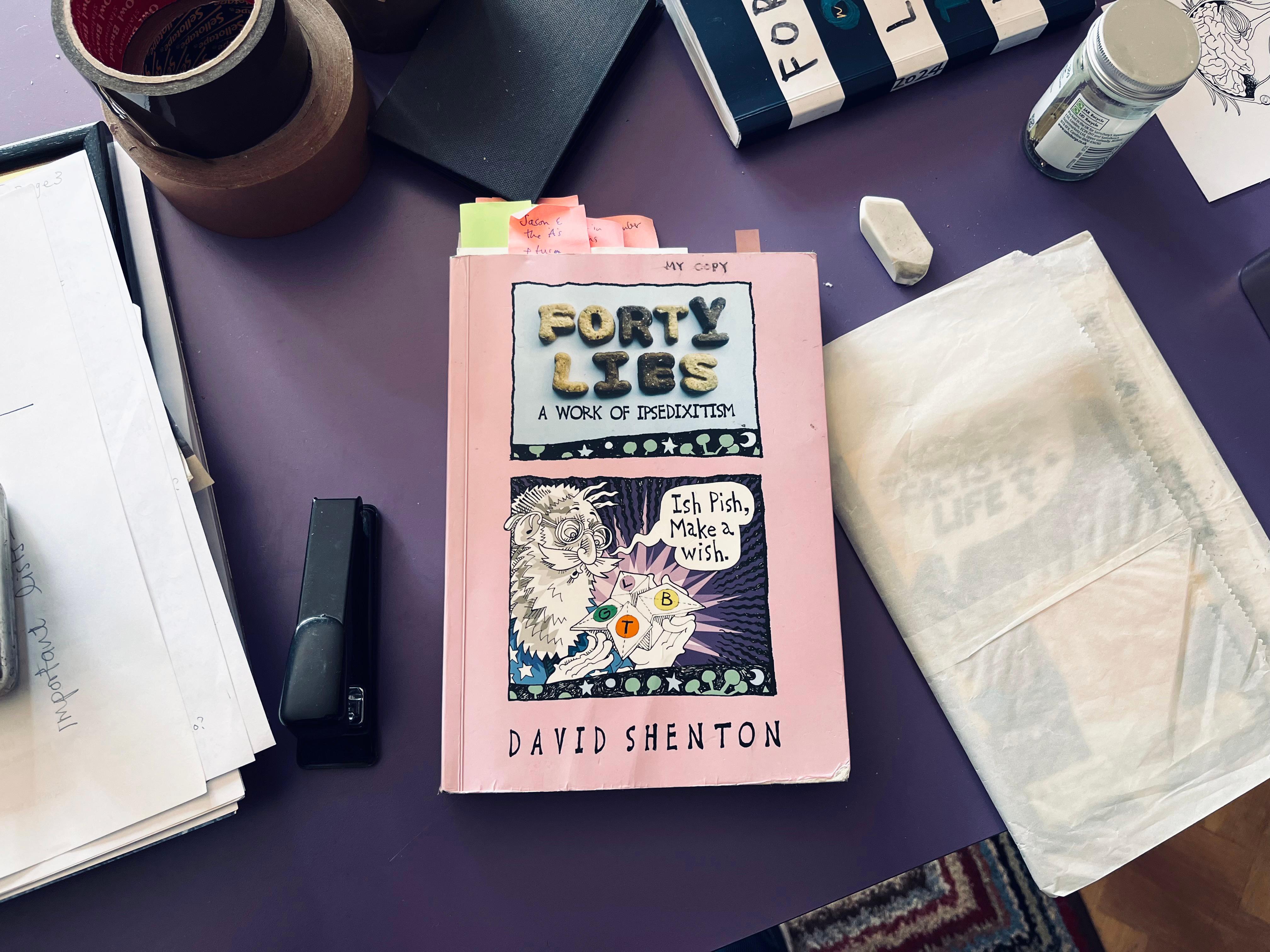 A desk showing oaoers and books. In the middle is the book 'Forty Lies' with post-its showing coming out the top.