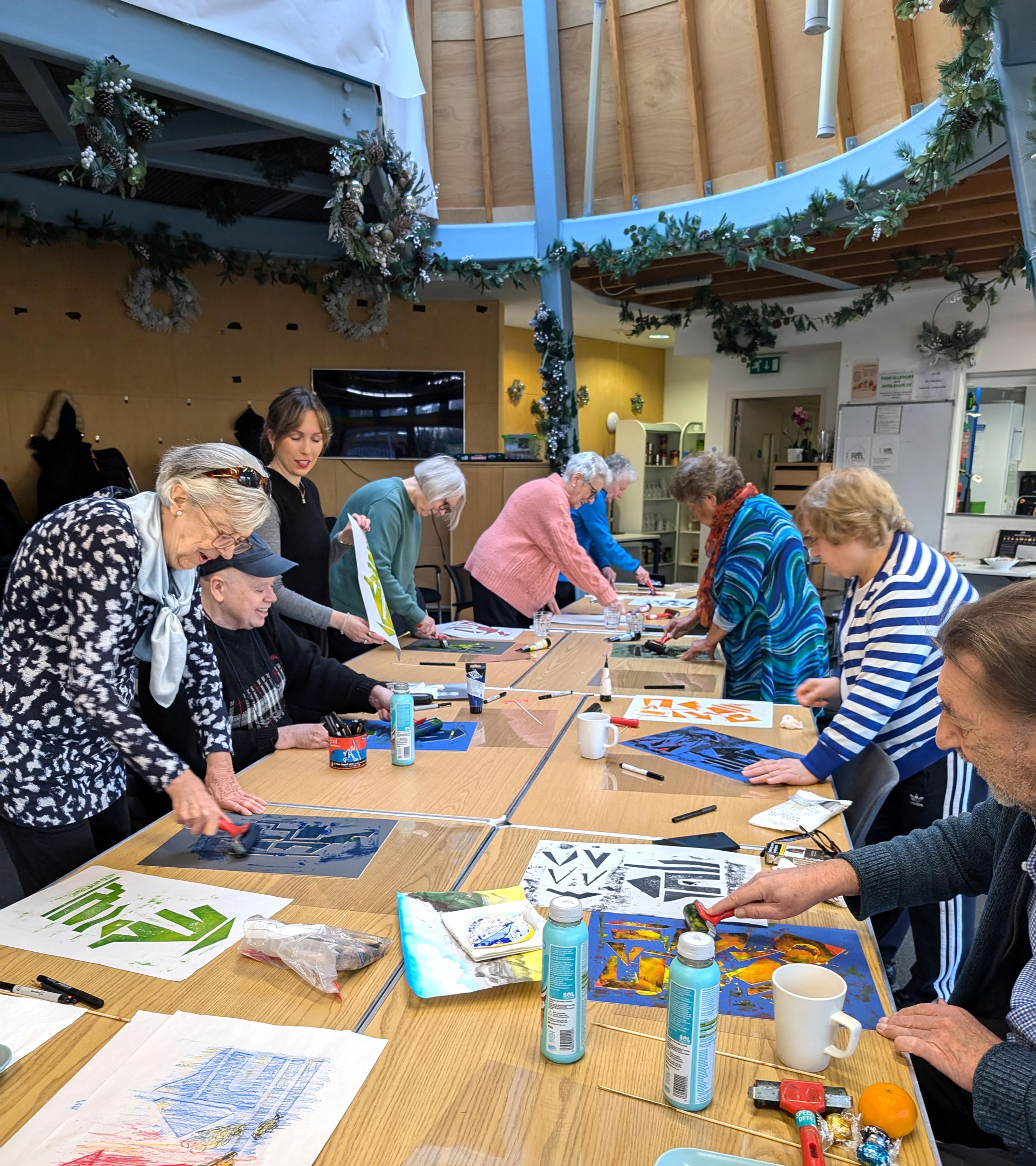 A photograph of a group of older adults gathered around a large table creating monoprints with ink, rollers and large sheets of paper