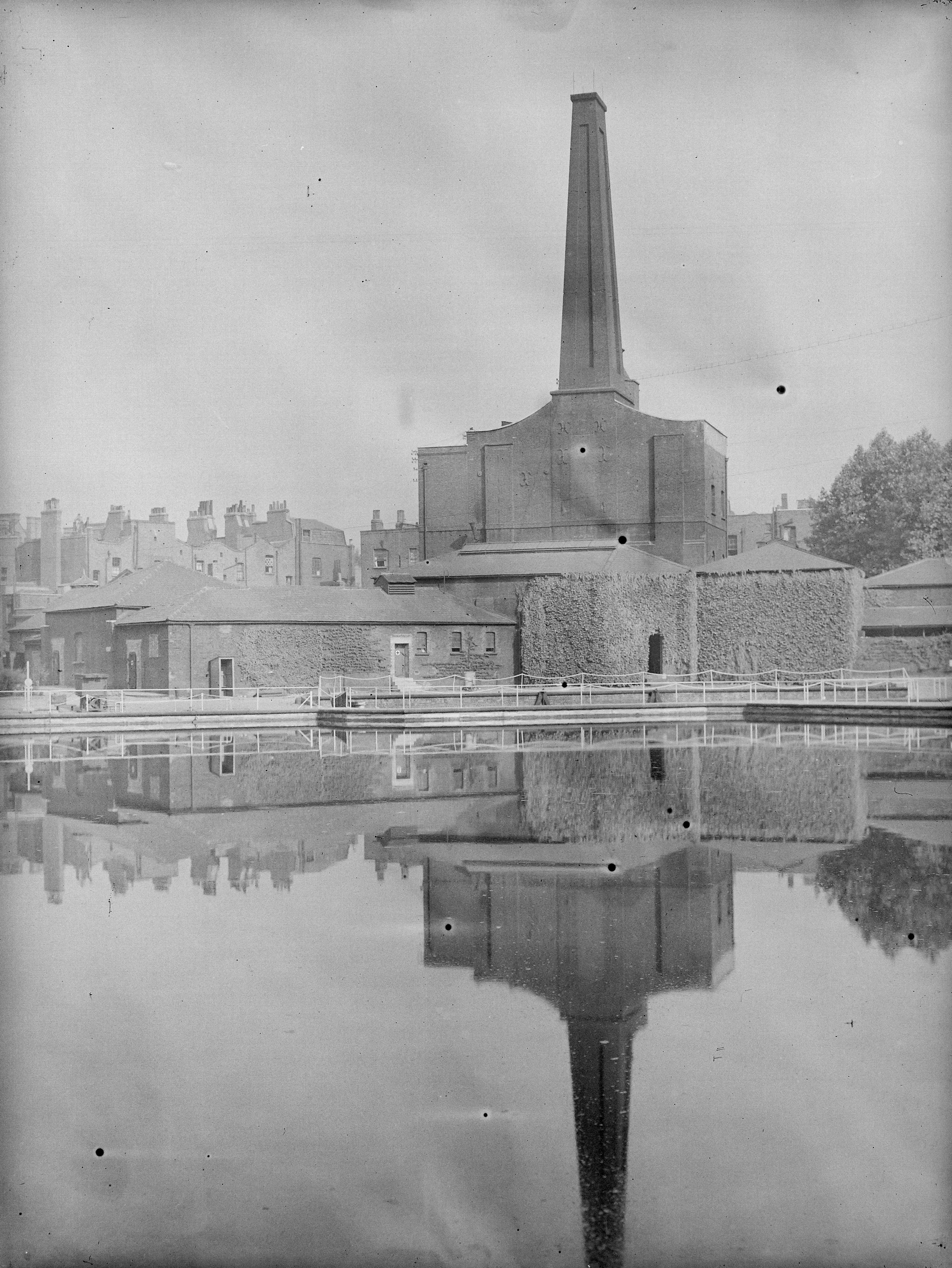 Black and white photograph of industrial brick buildings reflected in a pond in the foreground