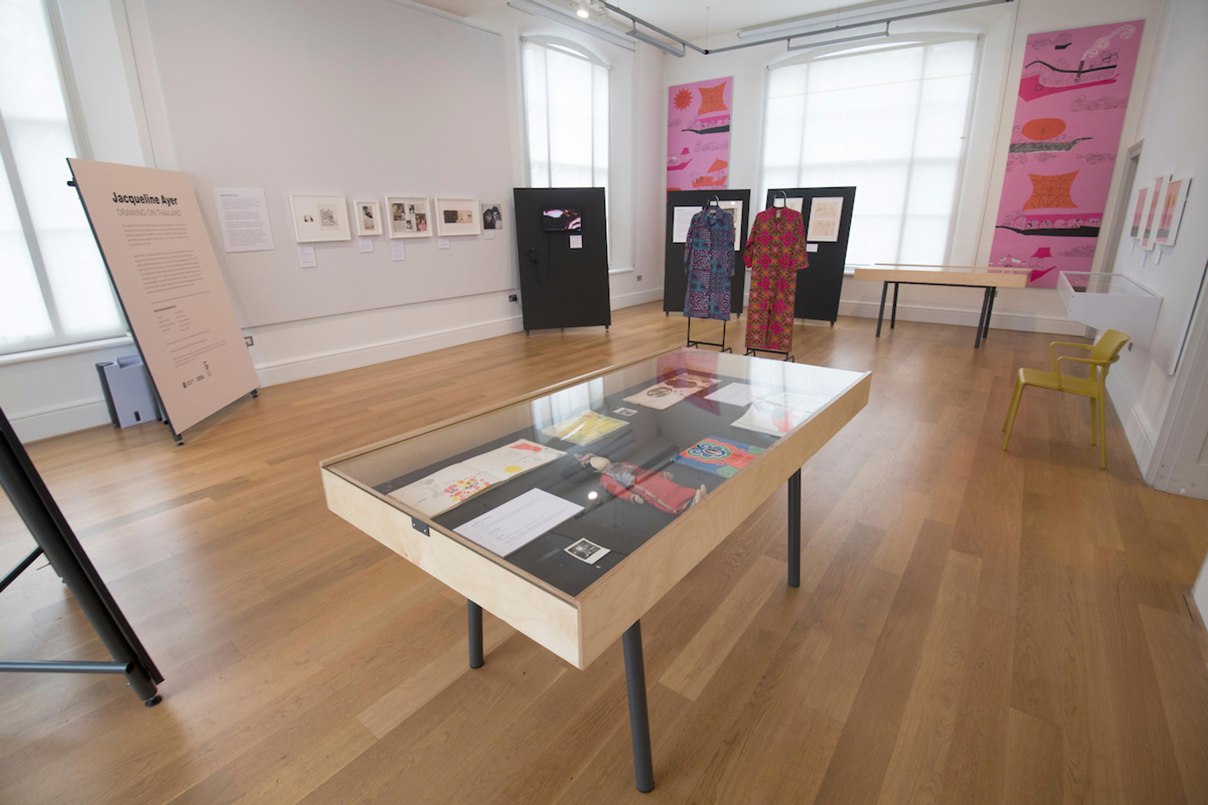 Inside an exhibition space with wall illustrations, framed illustrations on the walls, text displays and two glass table cabinets.