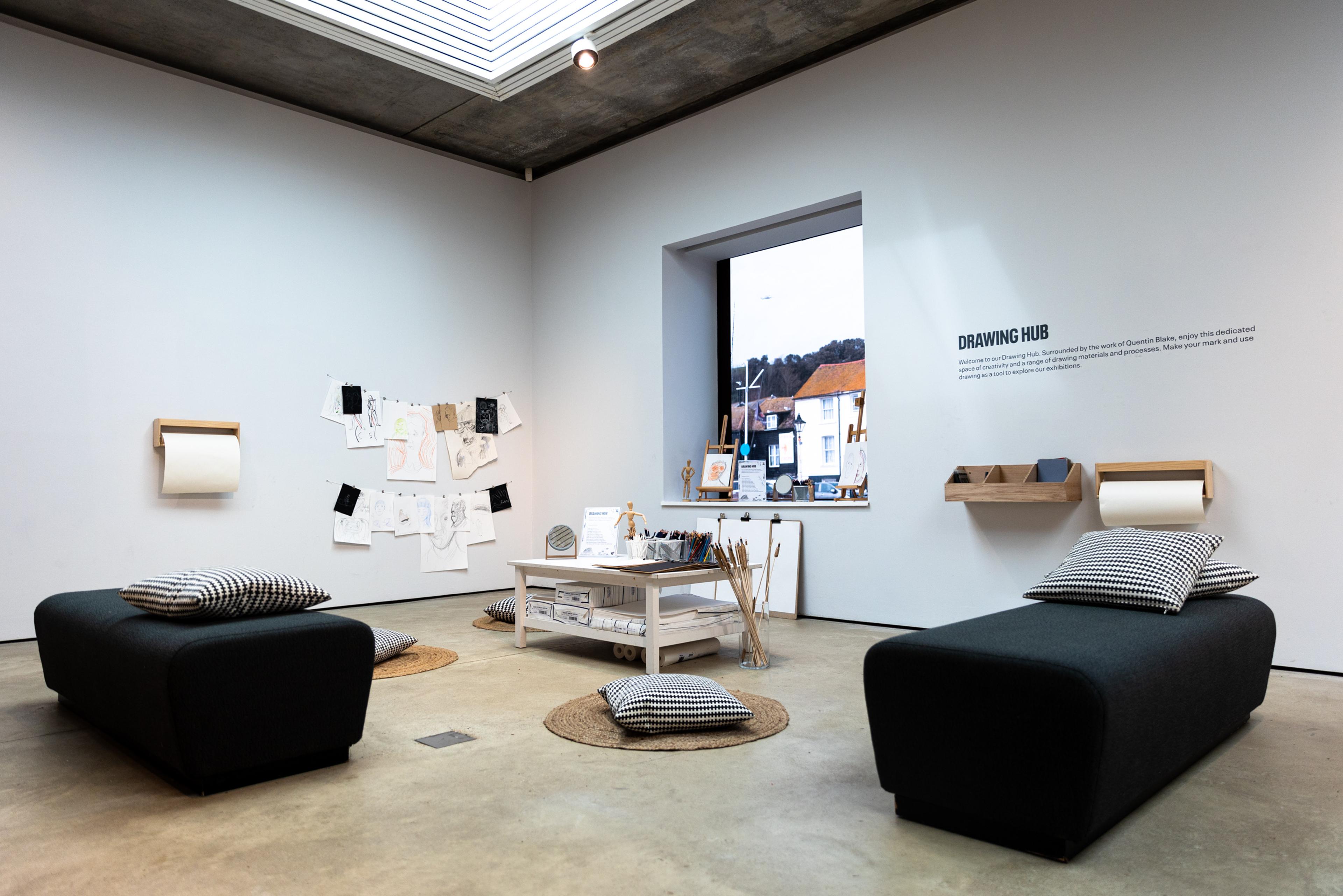 A creative space called the Drawing Hub with seating, cushions on the floor, a table in the middle of the space covered in art materials. Two paper dispensers are on the walls and illustrations are hung up on a string.