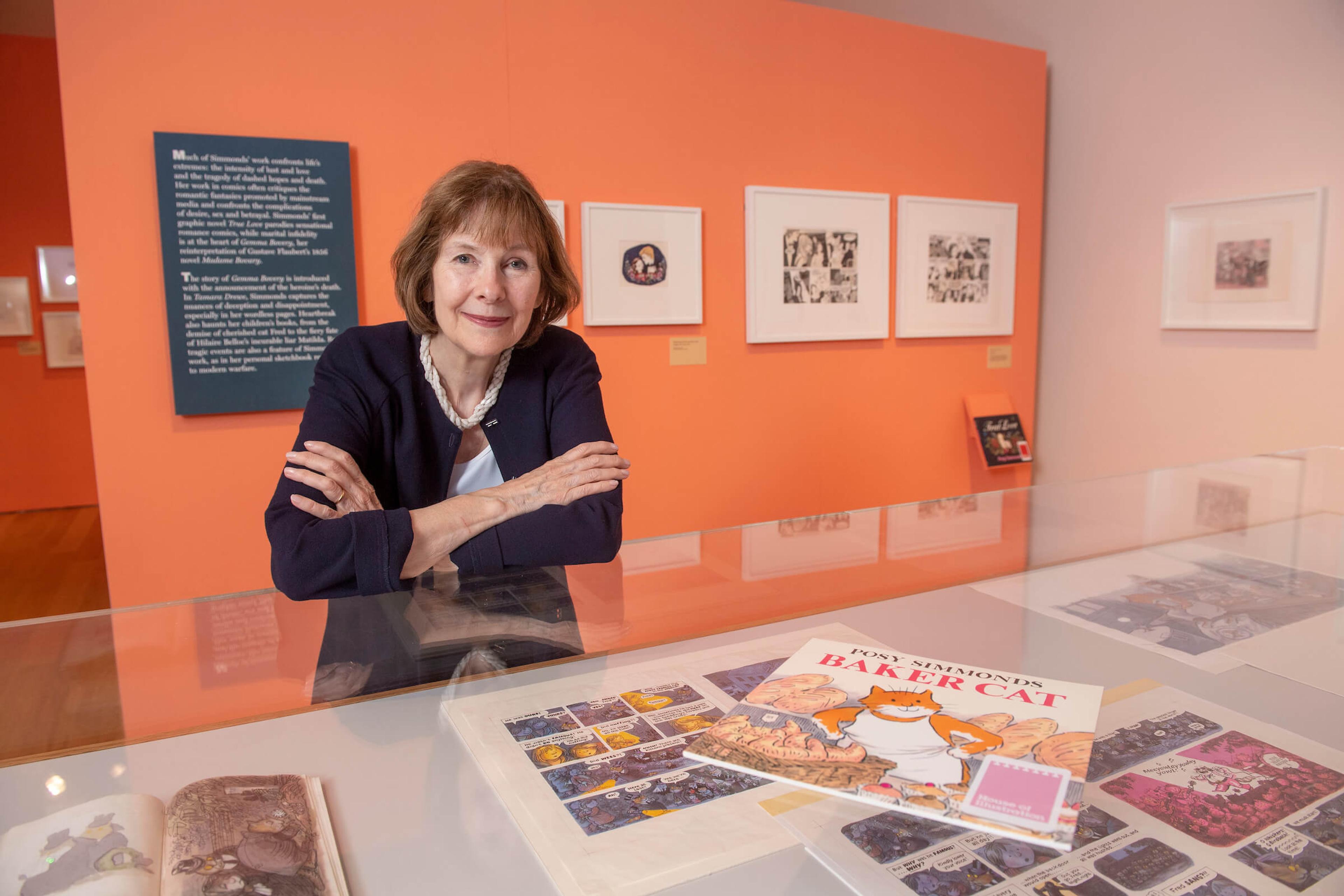 Posy Simmonds at the exhibition, standing behind a display case with pages from her comics