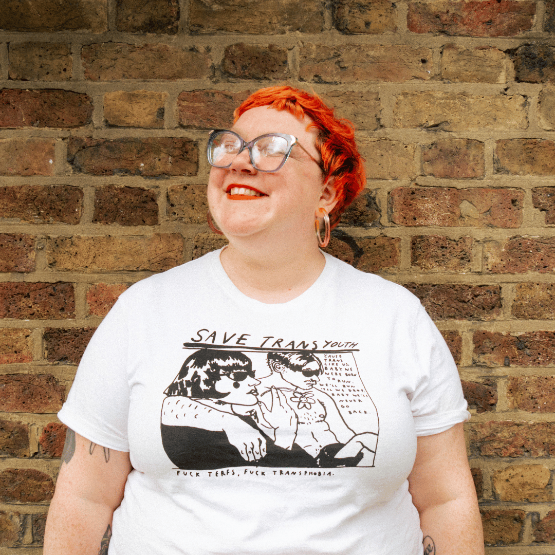 A woman with short red hair wearing a white t-shirt with black and white graphics stands against a brick background looking up and smiling.