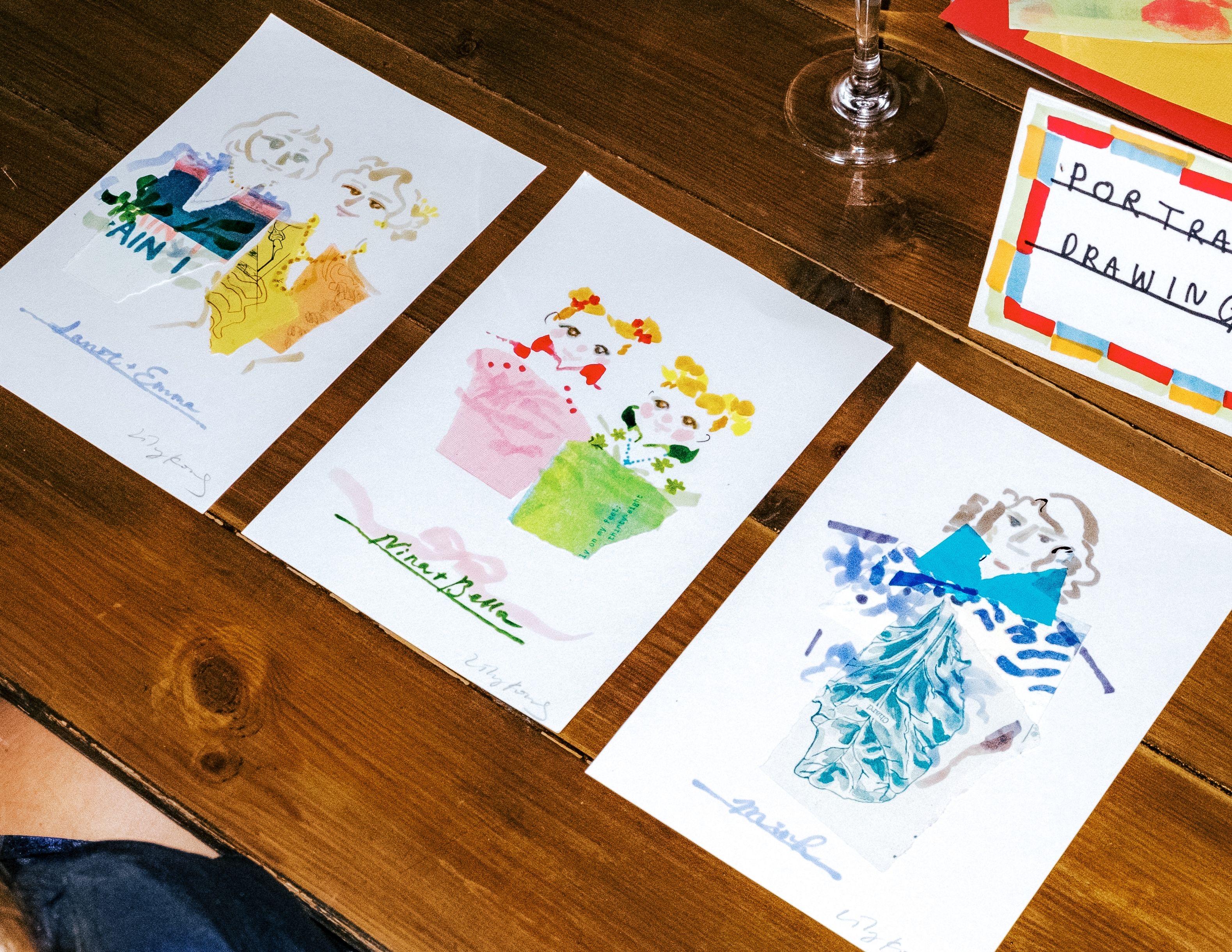 Three colourful portrait drawings sit lined up on a wooden table.