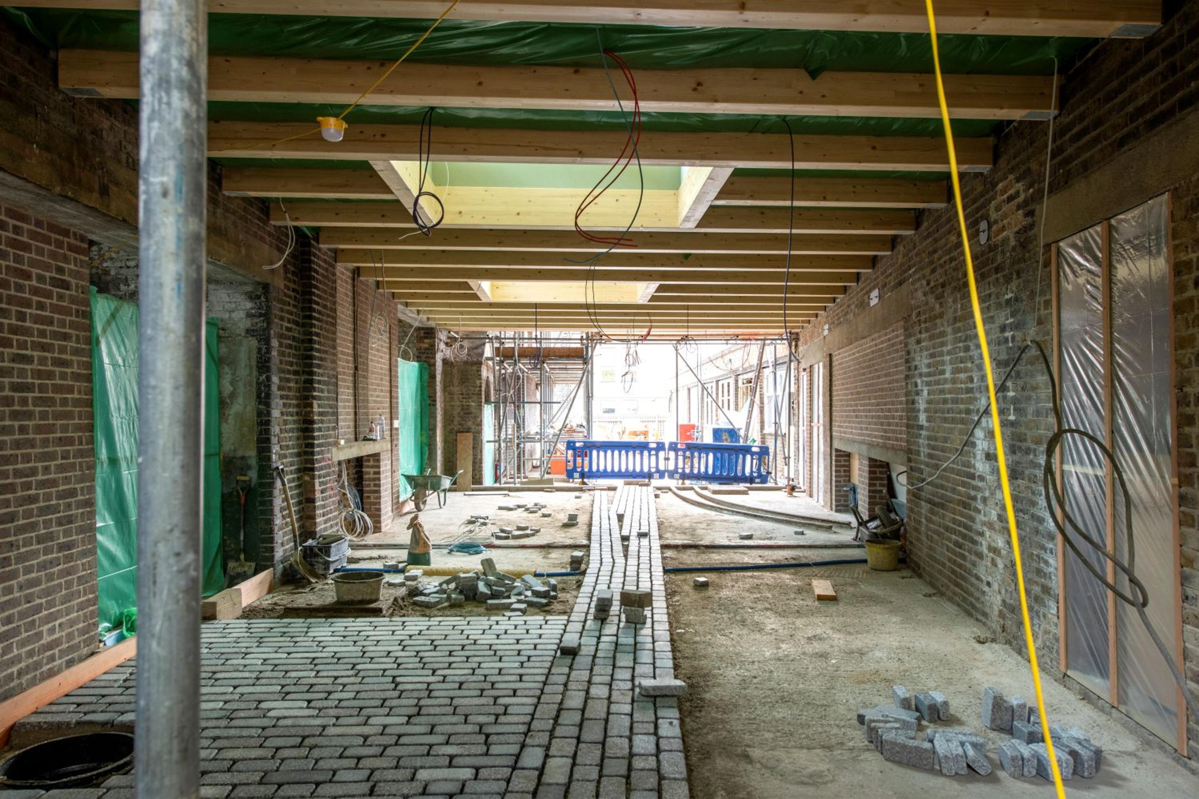 Construction site interior with partially built brick walls and an unfinished ceiling. Paved stone floor and light streaming from scaffolding outside.