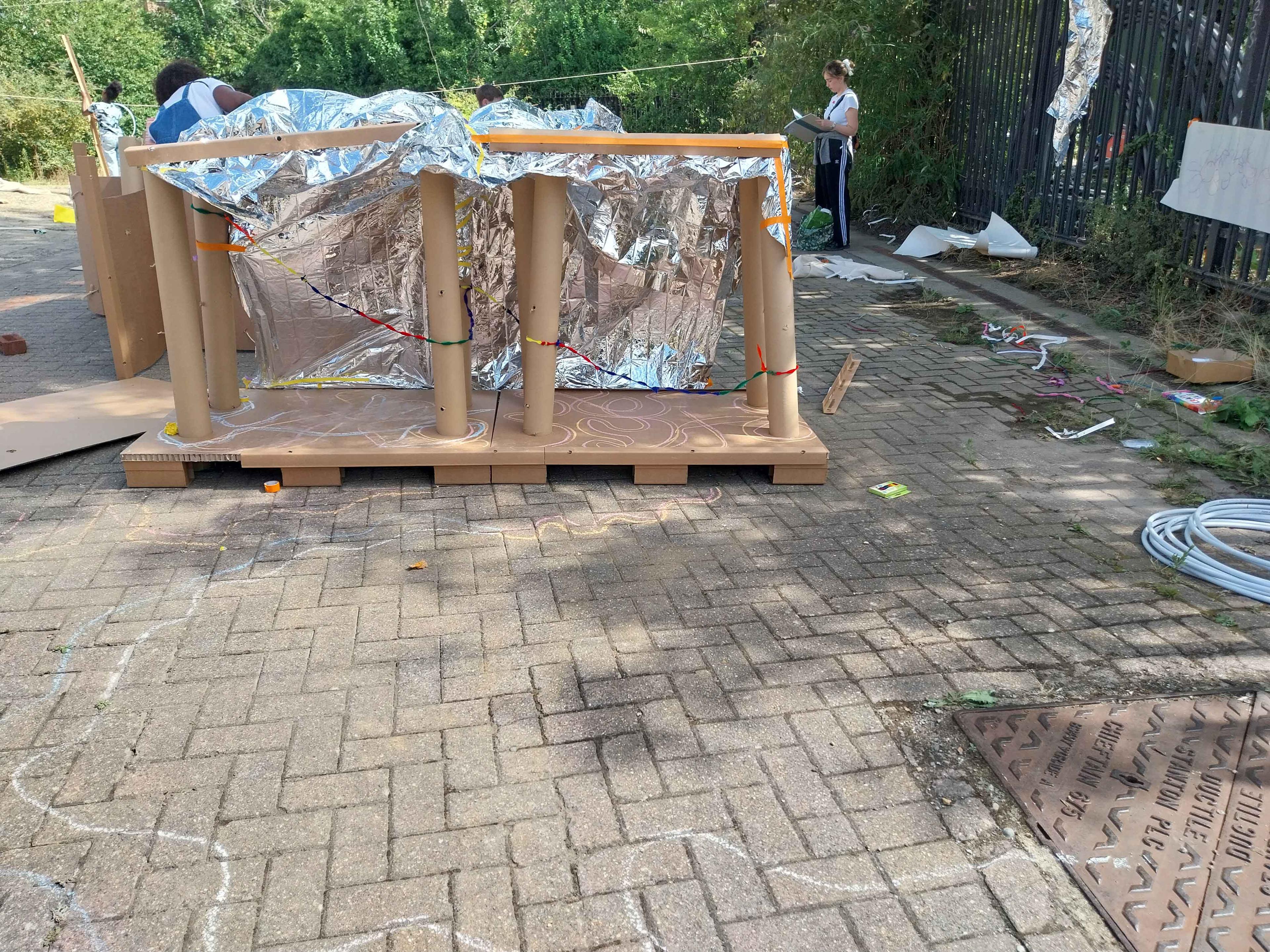 A person illustrating outside on a brick ground. A large wooden and foil sculpture is in front of them. There is chalk drawings on the ground. 