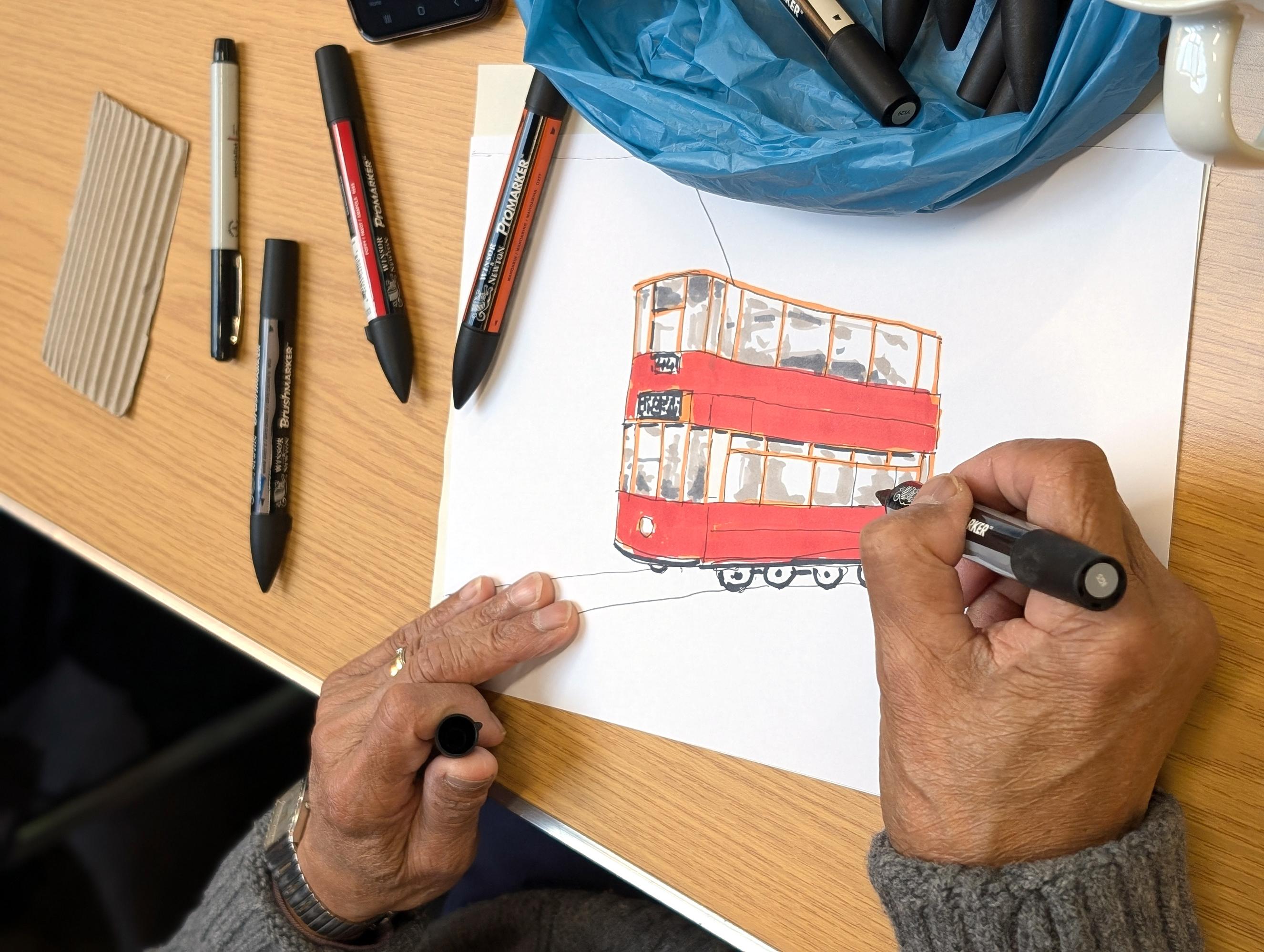An illustration of a red tram, executed in coloured felt pen. An adult's hand is holding a black pen and adding details to the drawing.