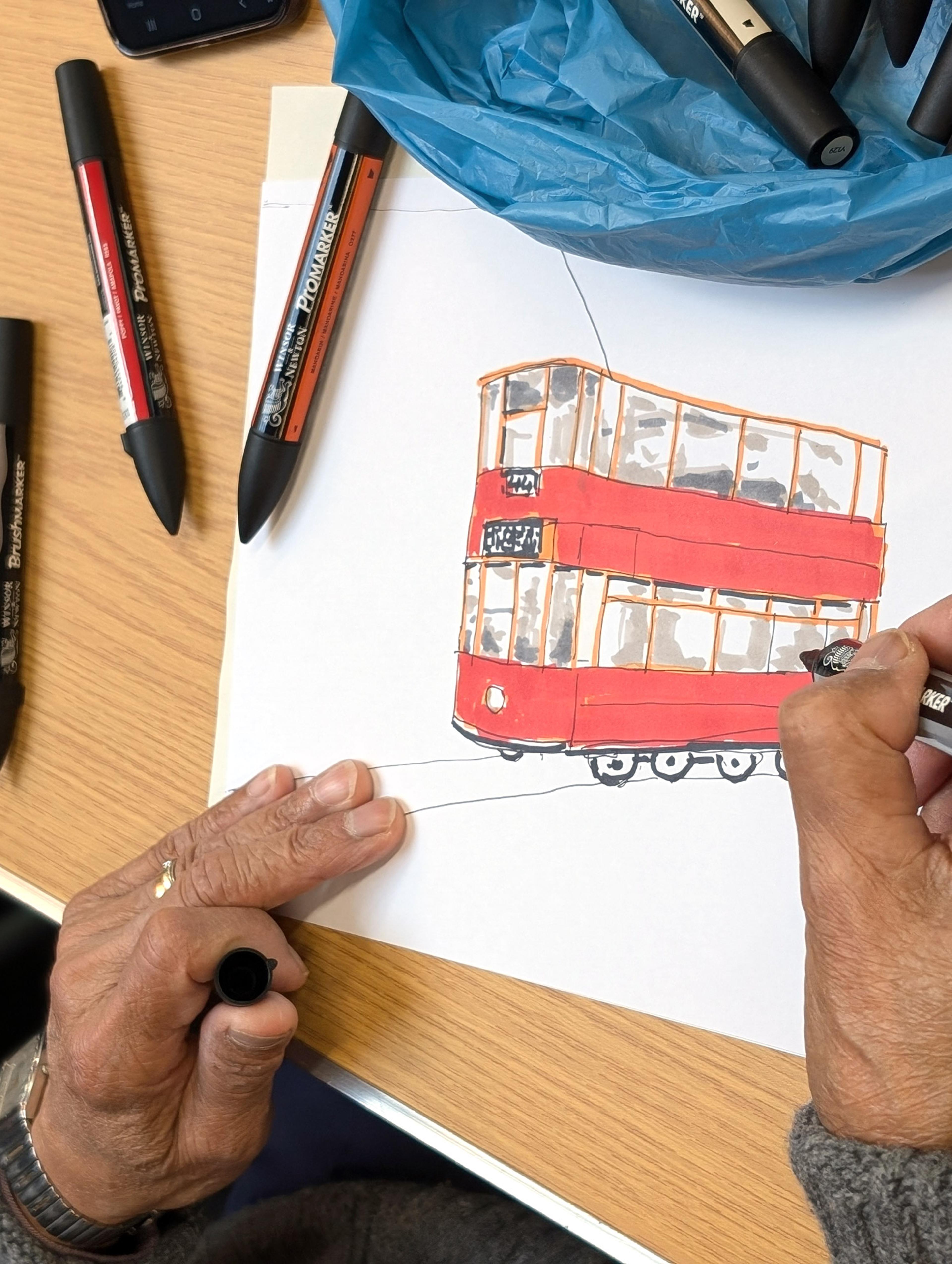 An illustration of a red tram, executed in coloured felt pen. An adult's hand is holding a black pen and adding details to the drawing.