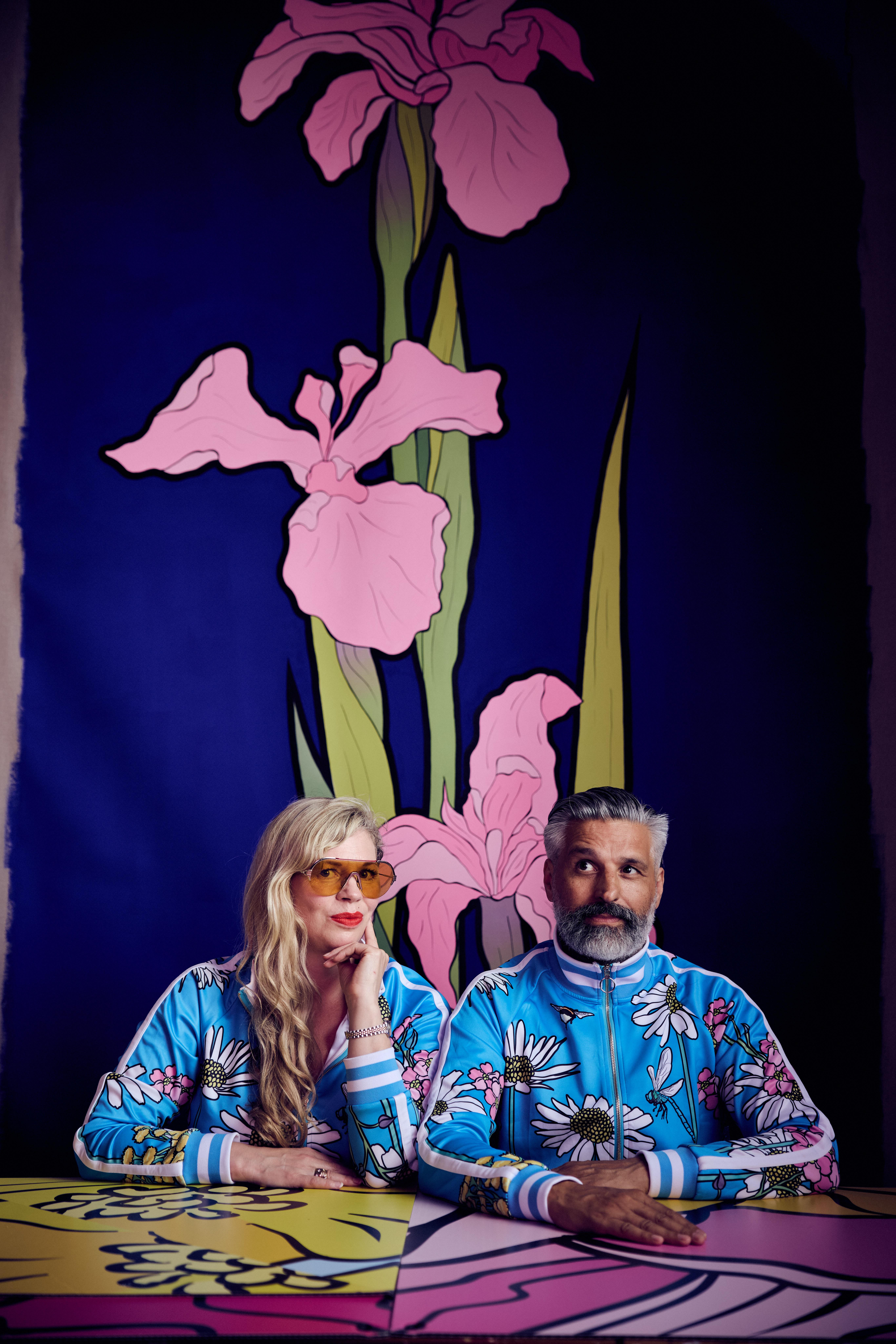 Two artists in matching floral tracksuit tops look out to the camera, with a floral design in the background.