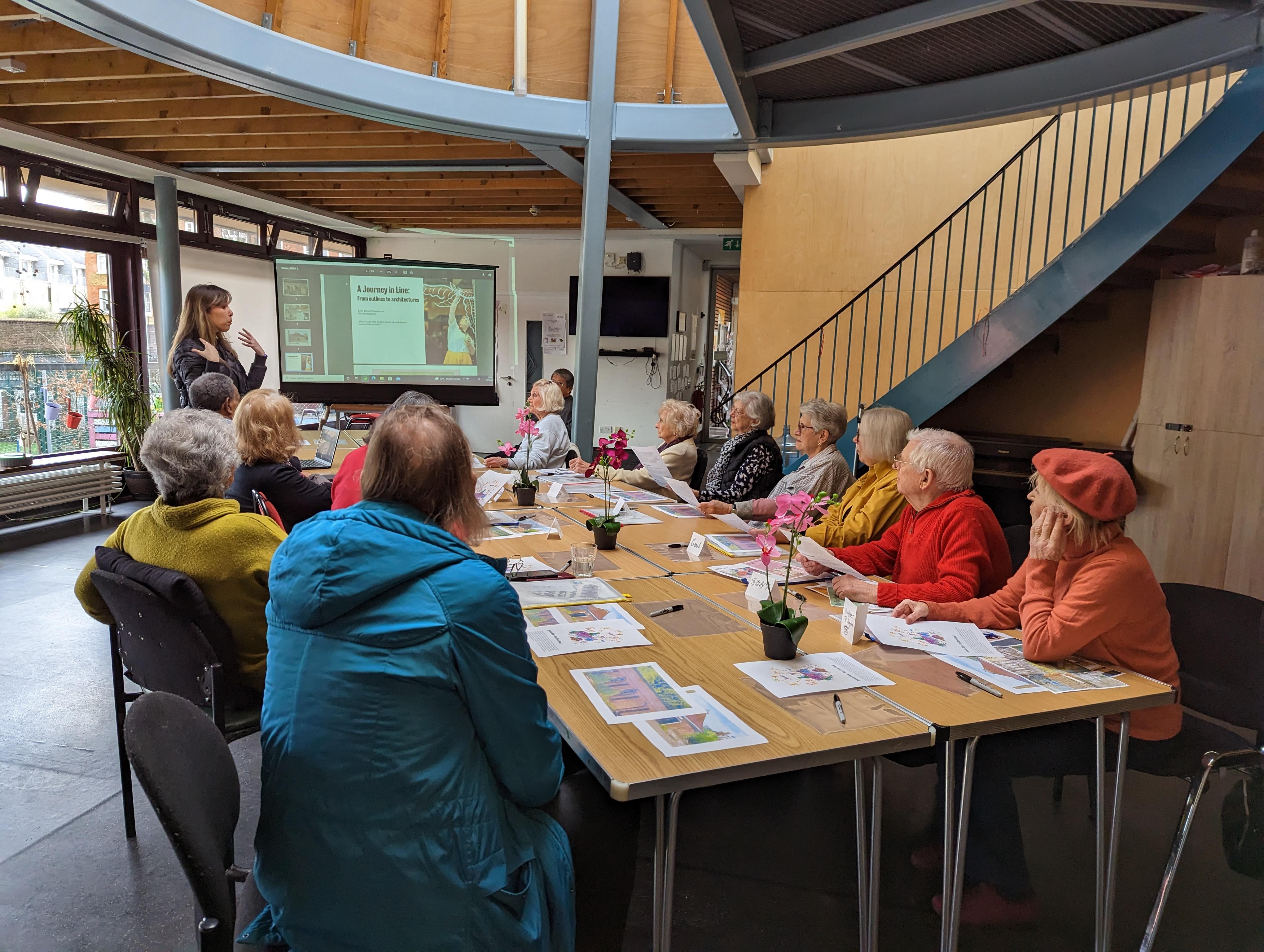 A group of older people sat at a table, seen from behind. They're looking at the workshop facilitator who appears to be addressing them. On the table, we see sheets of paper and some ink pots with art material.