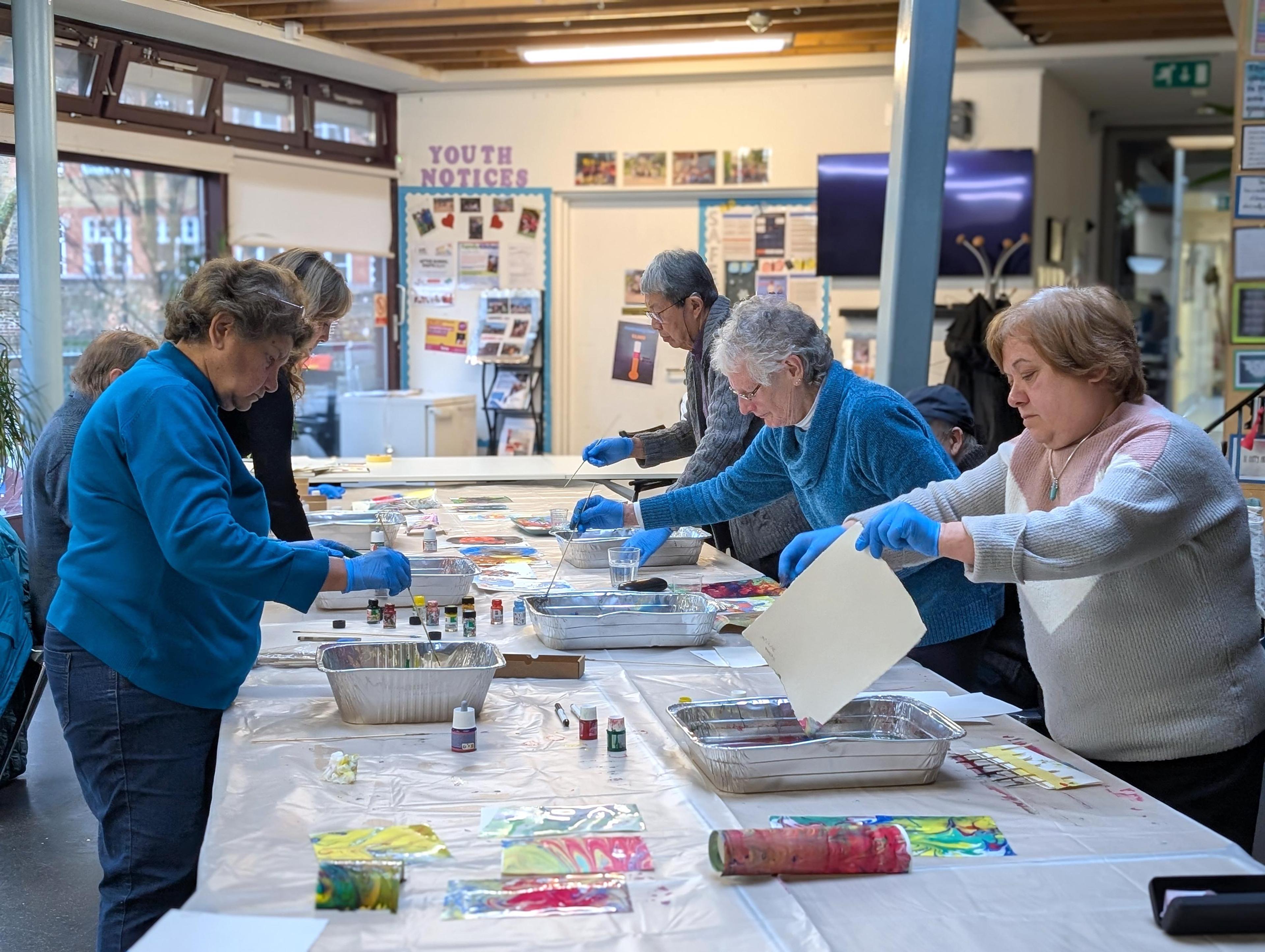 A group of older adults stood around a large table. They are dipping sheets of paper into trays of paint to create a marbled effect. 