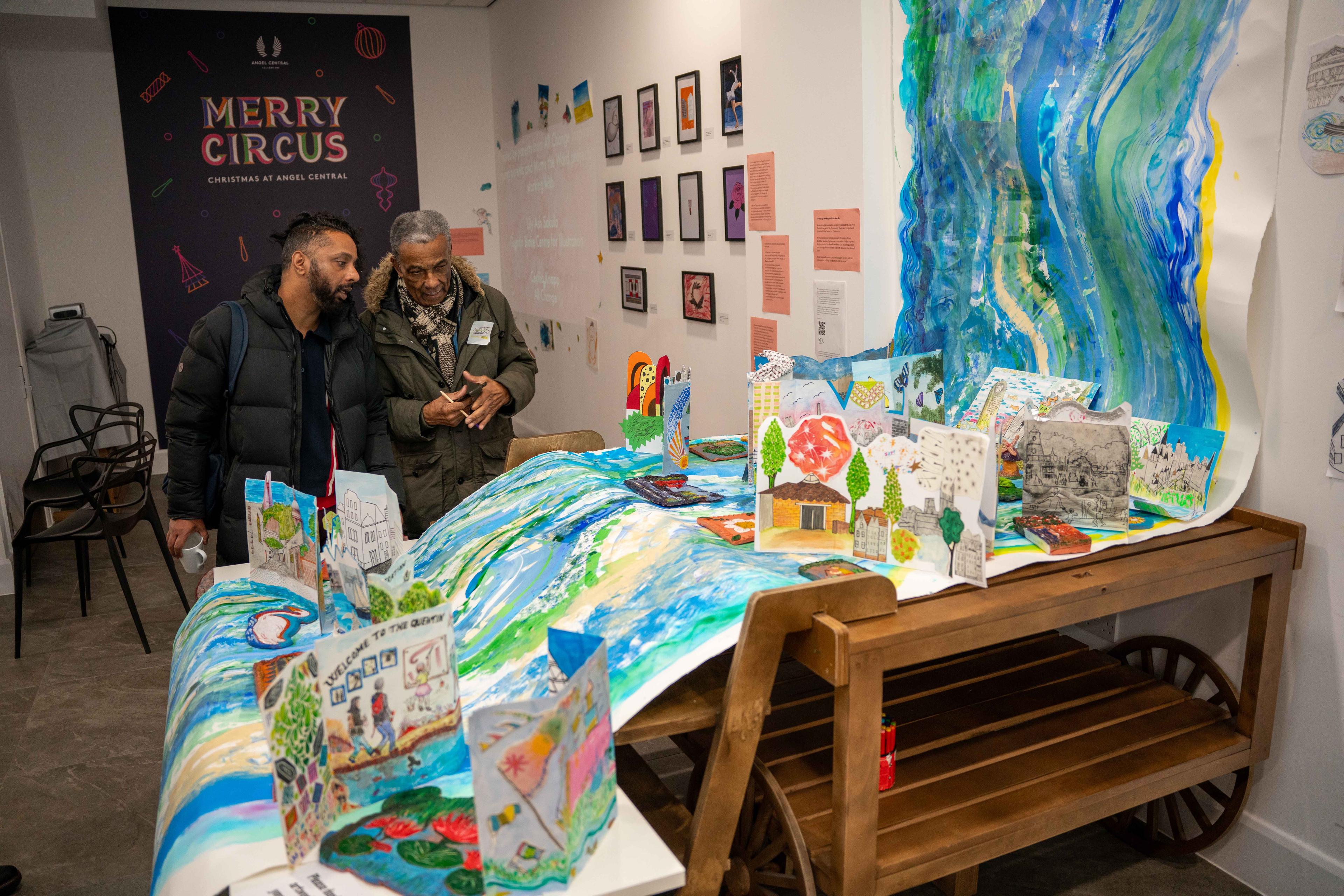 Two people stand next to an installation of a large paper scroll with a river painted onto it, this hangs from a wooden structure and to the floor. There are paper and ceramic artworks on top of the paper scroll at certain points.