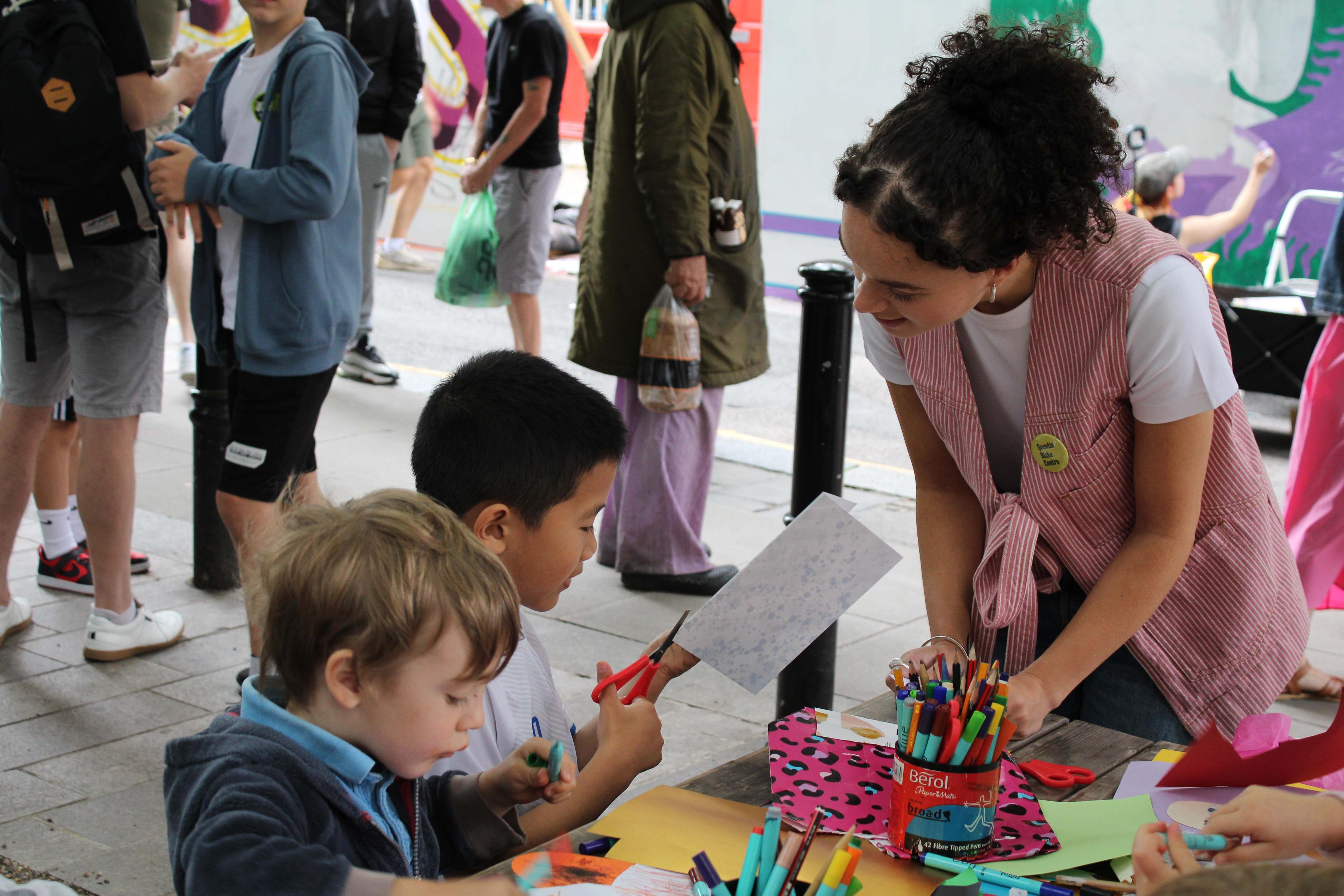 A woman talking to two young children who are drawing and cutting paper with scissors