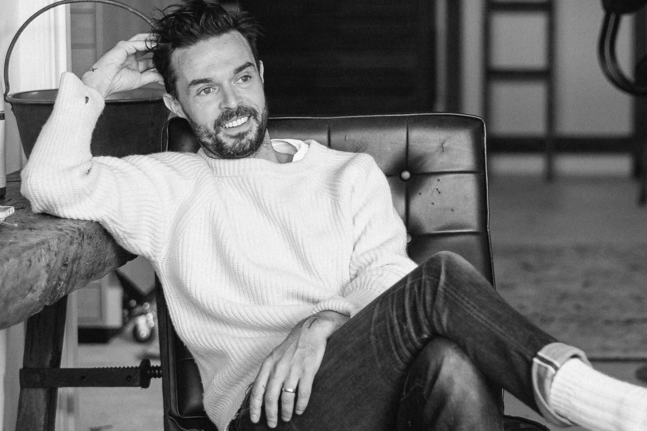 A man sits smiling in a chair resting his head on his head