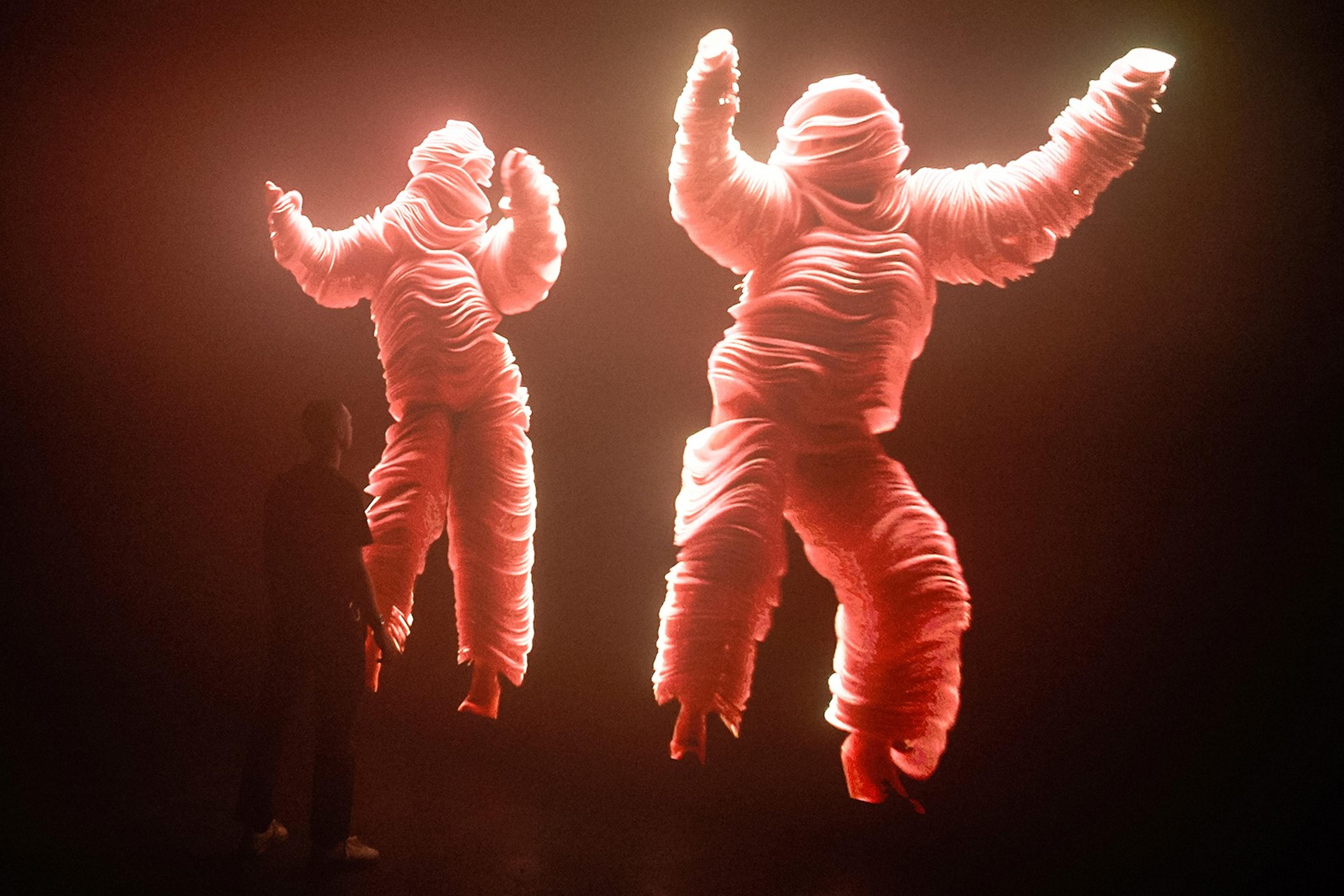 A photograph of a person standing in front of a screen in a gallery. The screen shows a large brightly lit image of two figures dancing in pink costumes on a black background