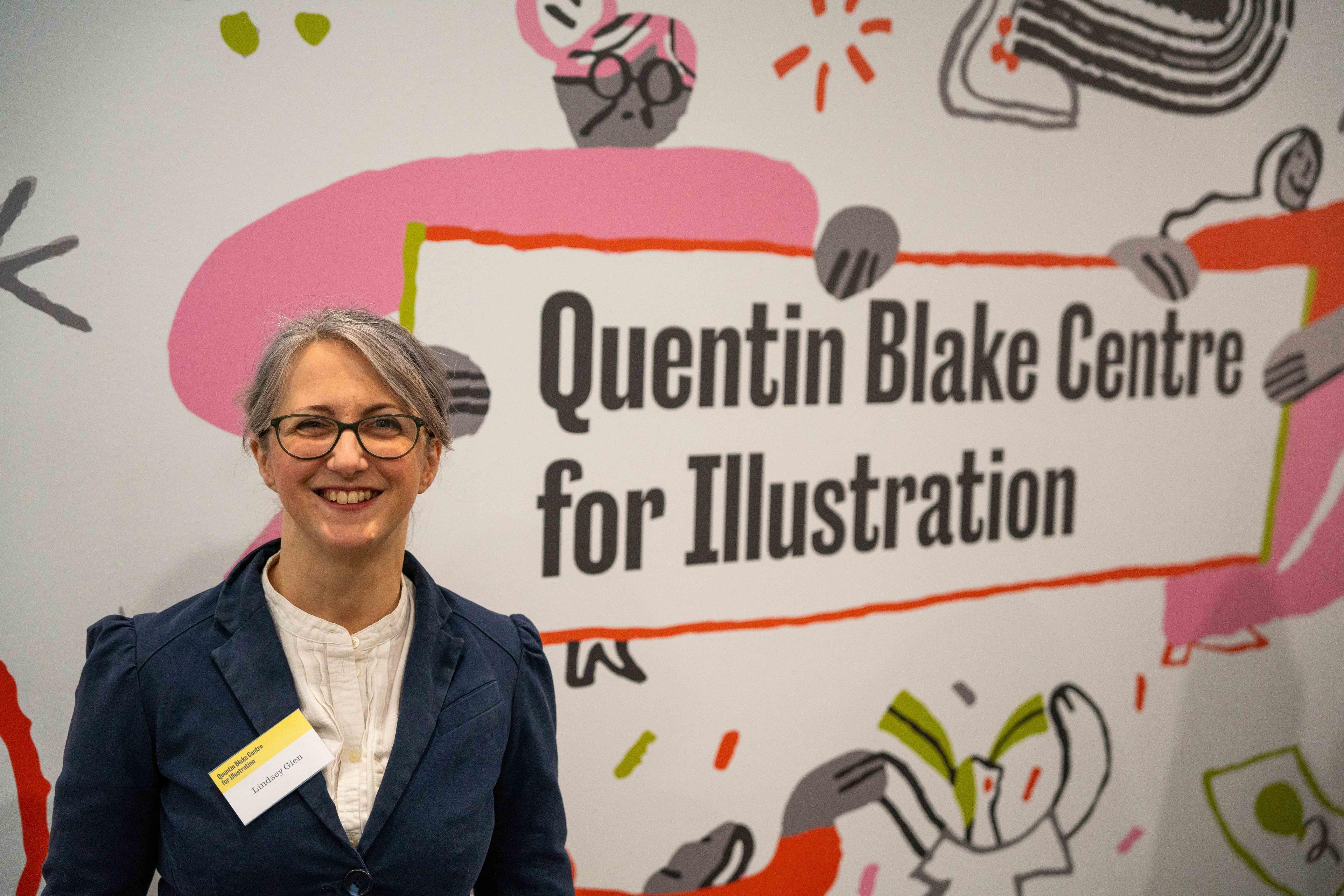 Photograph of a person standing and smiling in front of an illustrated wall with the wording 'Quentin Blake Centre for Illustration'.