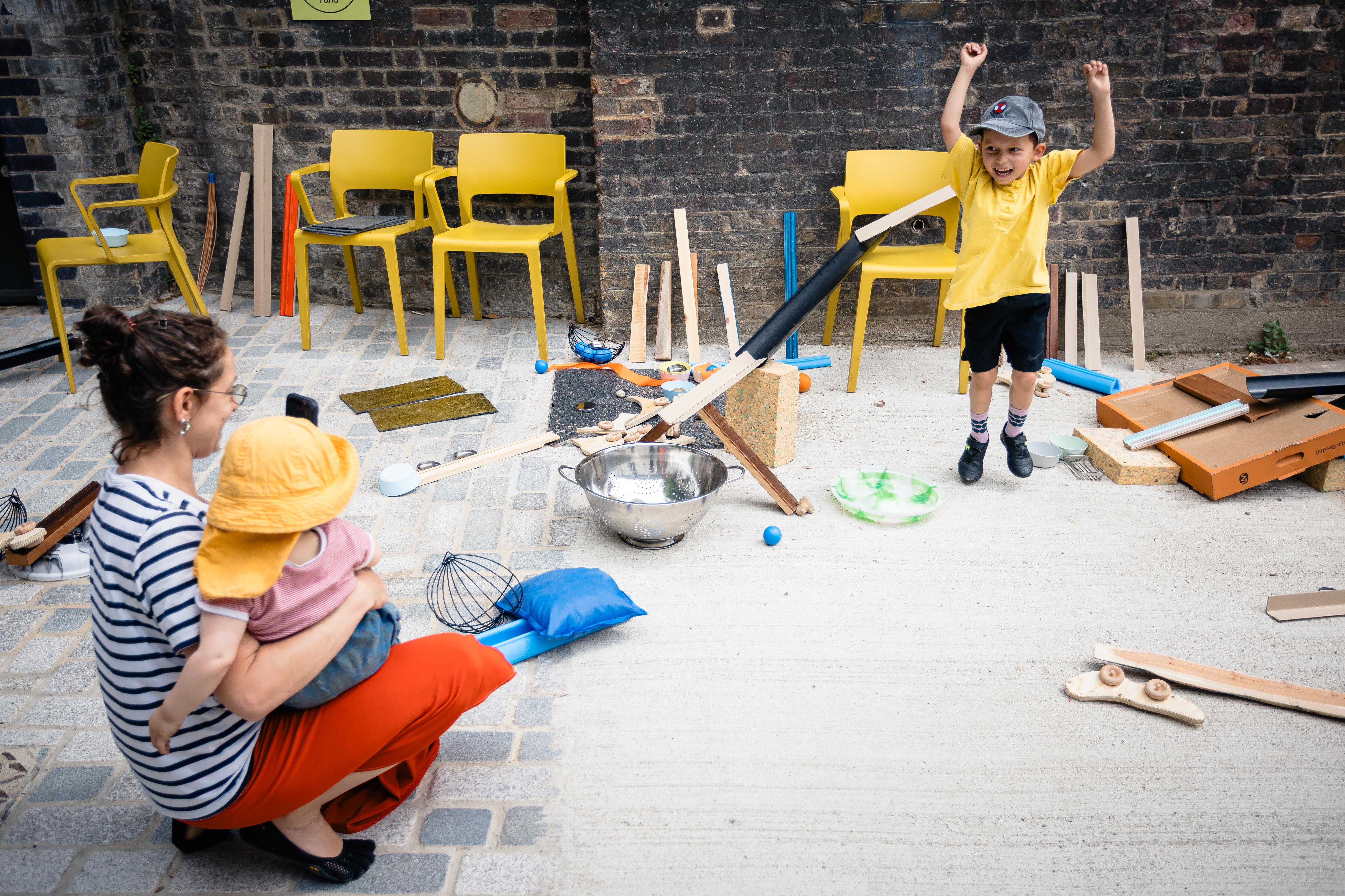 A woman sits with a child on her leg while a child celebrates the success of a track created from guttering and other everyday objects.