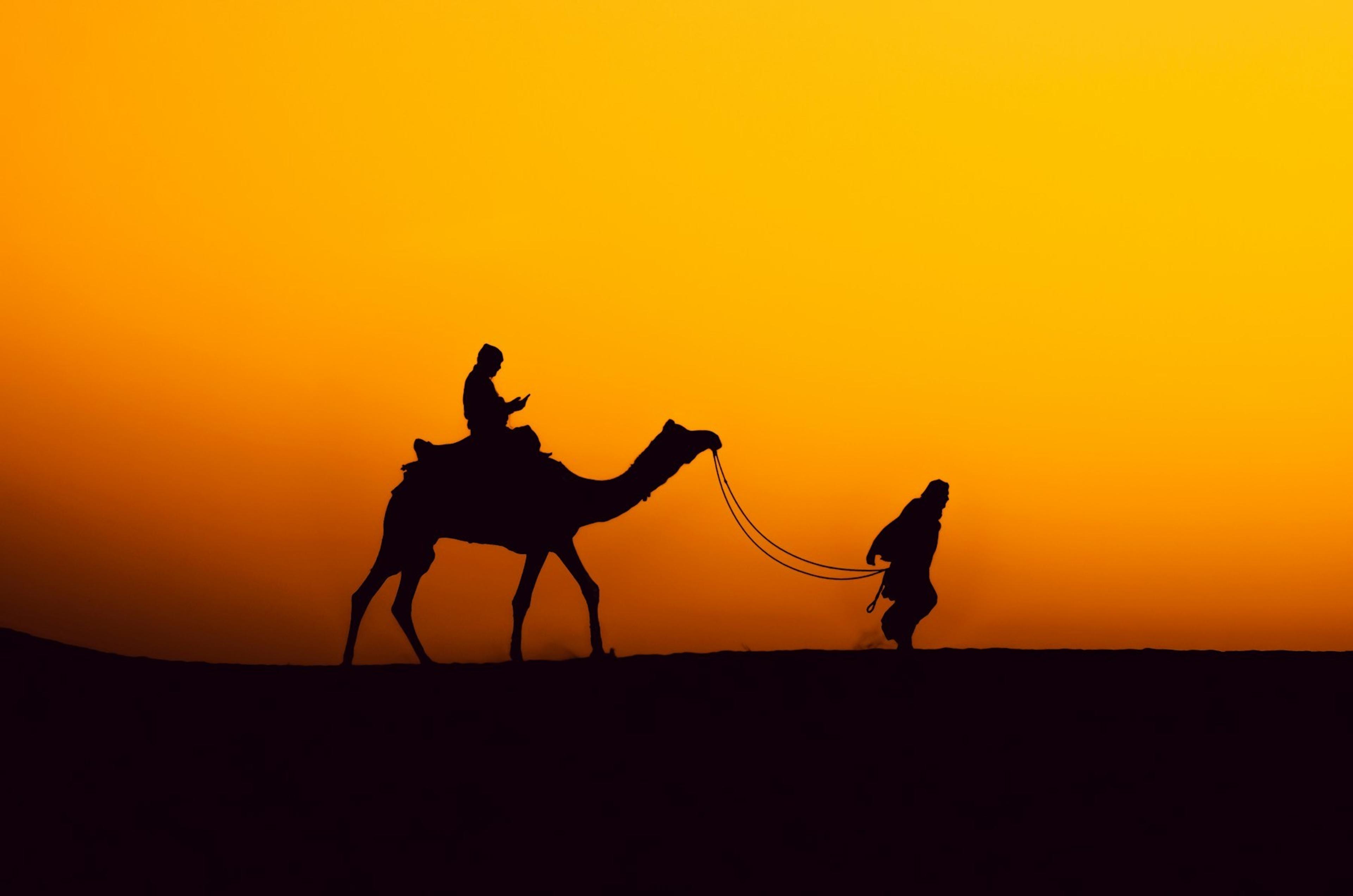 A camel and its handler silhouetted against an orange sunset on the sand dunes of Jaisalmer