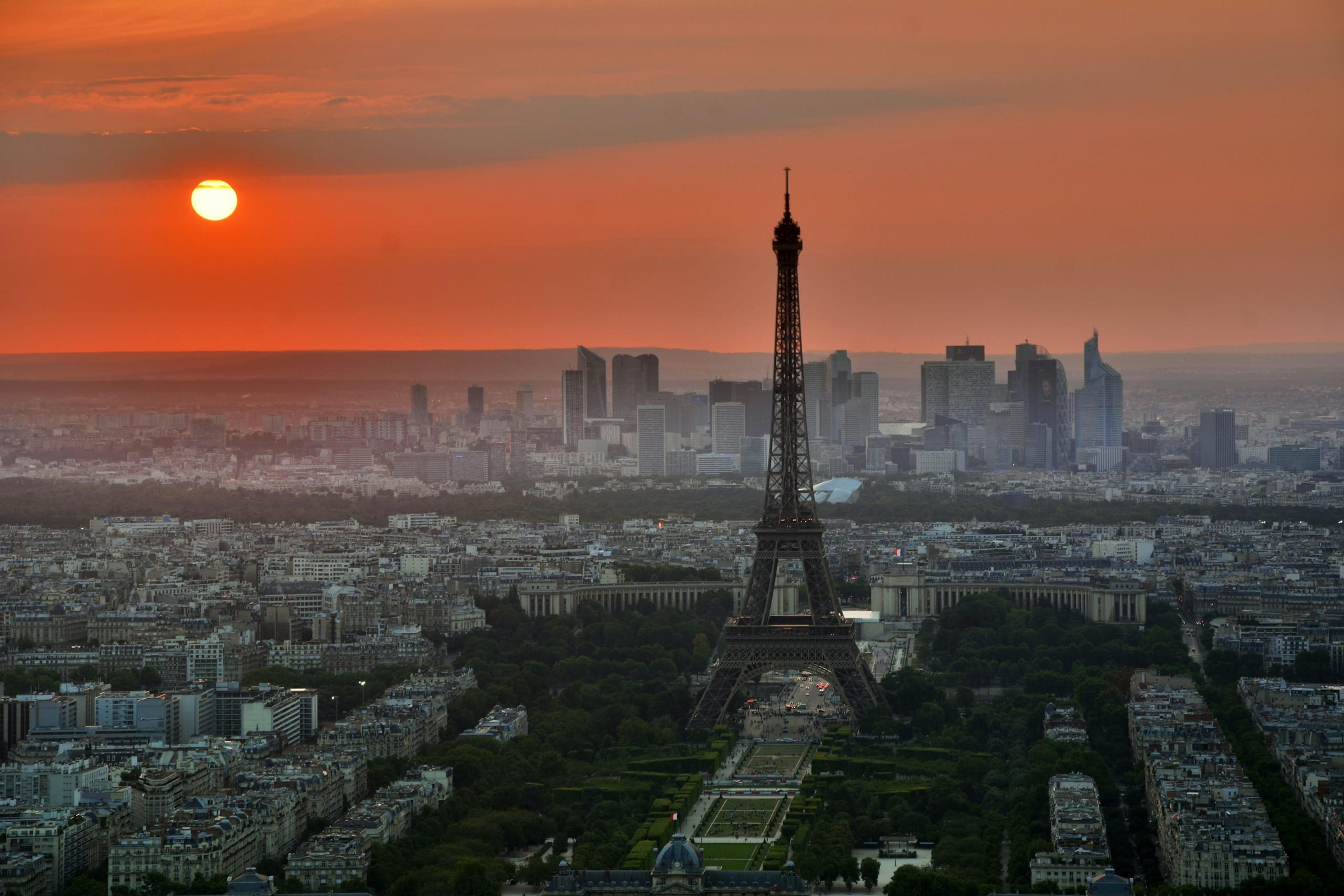The Eiffel Tower silhouetted against an orange sunset over the Paris skyline, with La Défense towers on the horizon.
