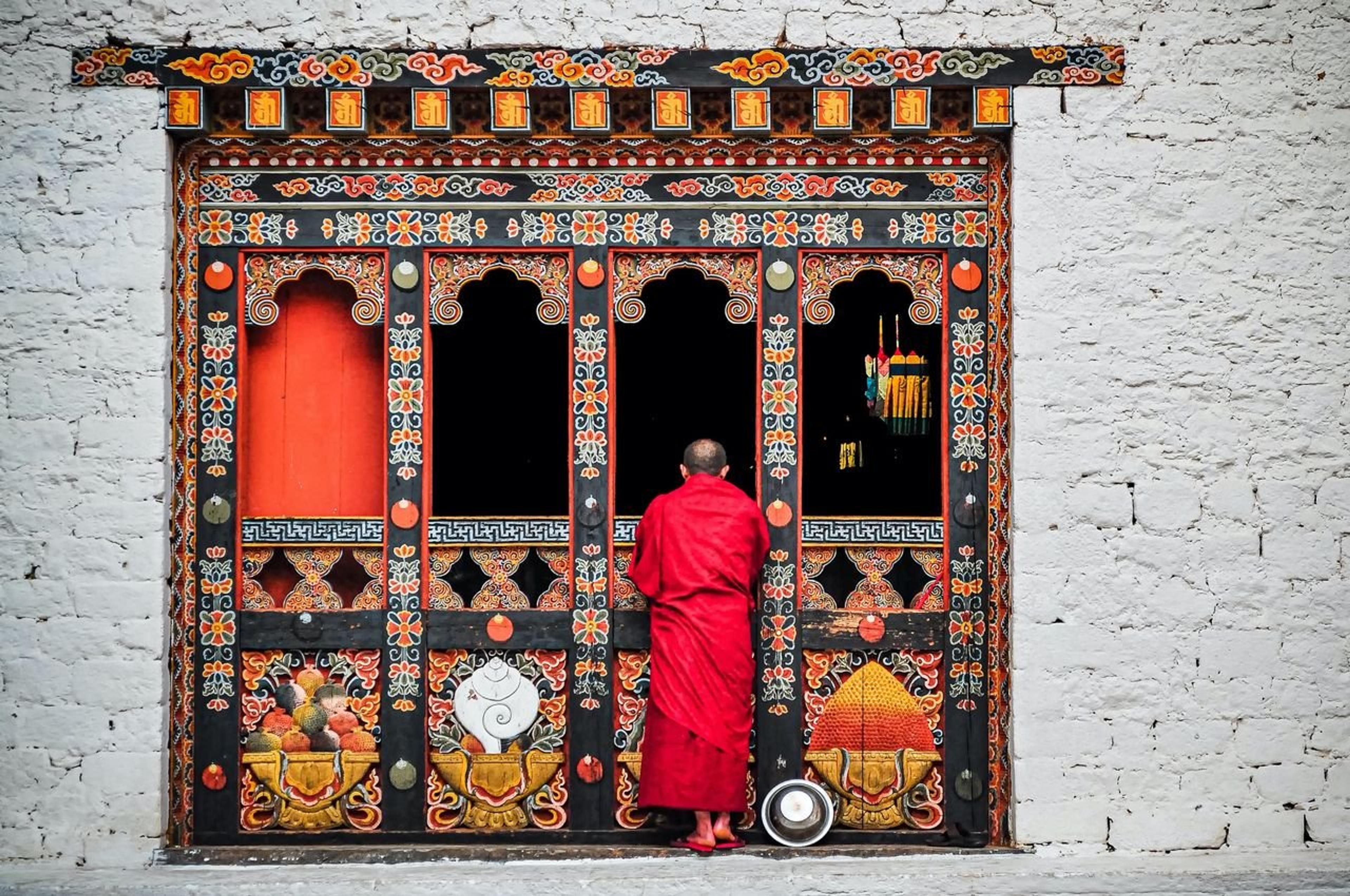 A Buddhist monk in red robes standing at an ornately decorated temple doorway in Bhutan.
