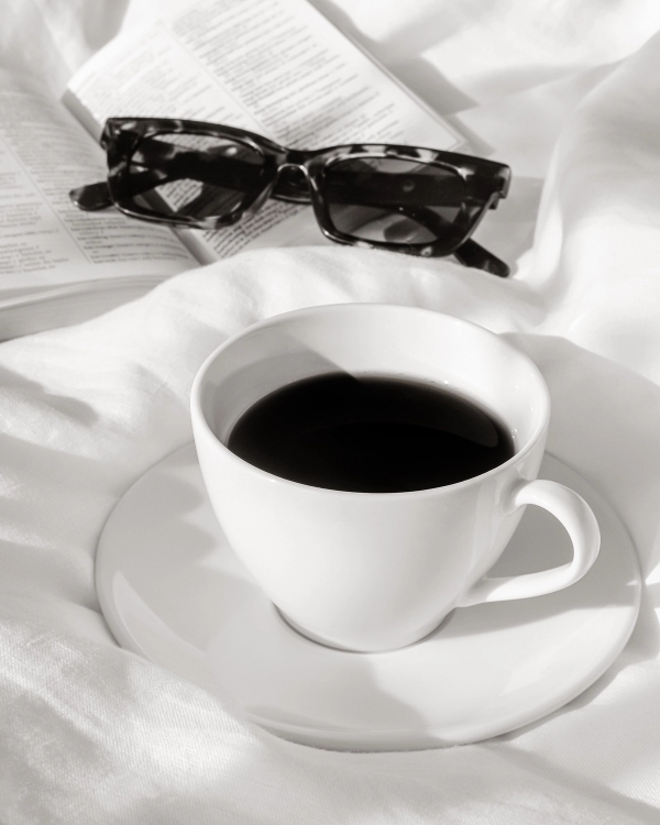 Black and white photo of a white coffee cup on bed linen with reading glasses and an open book