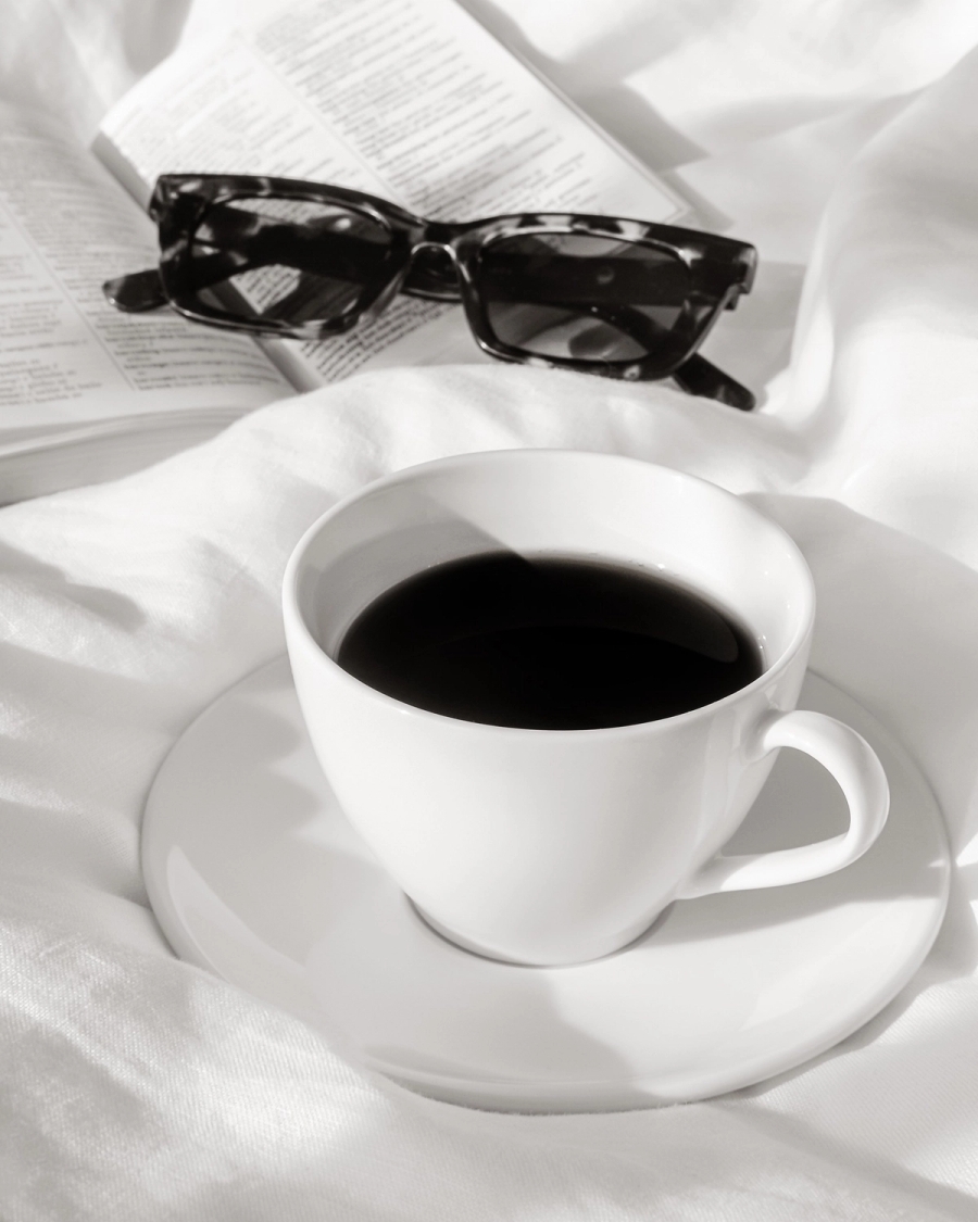 Black and white photo of a white coffee cup on bed linen with reading glasses and an open book