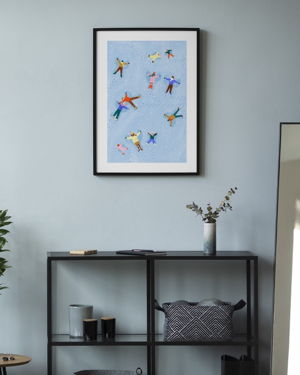 Angels in the Snow poster in a slim black frame on a pale blue wall above a black metal console shelf