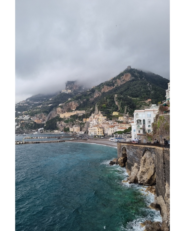 Colorful cliffside town overlooking turquoise sea under cloudy sky.