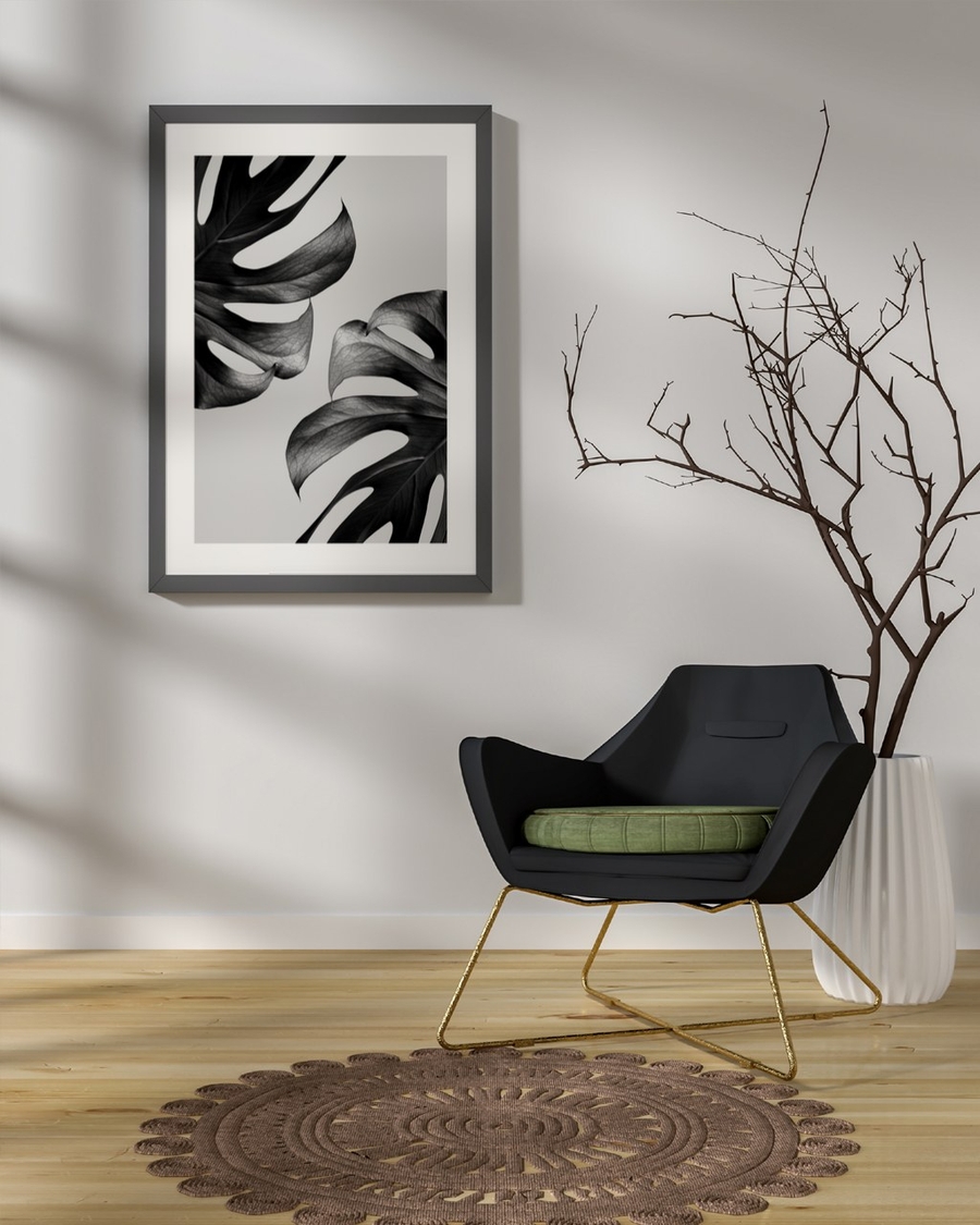 Monstera Noir poster in a dark grey frame above a black armchair in a bright minimal room
