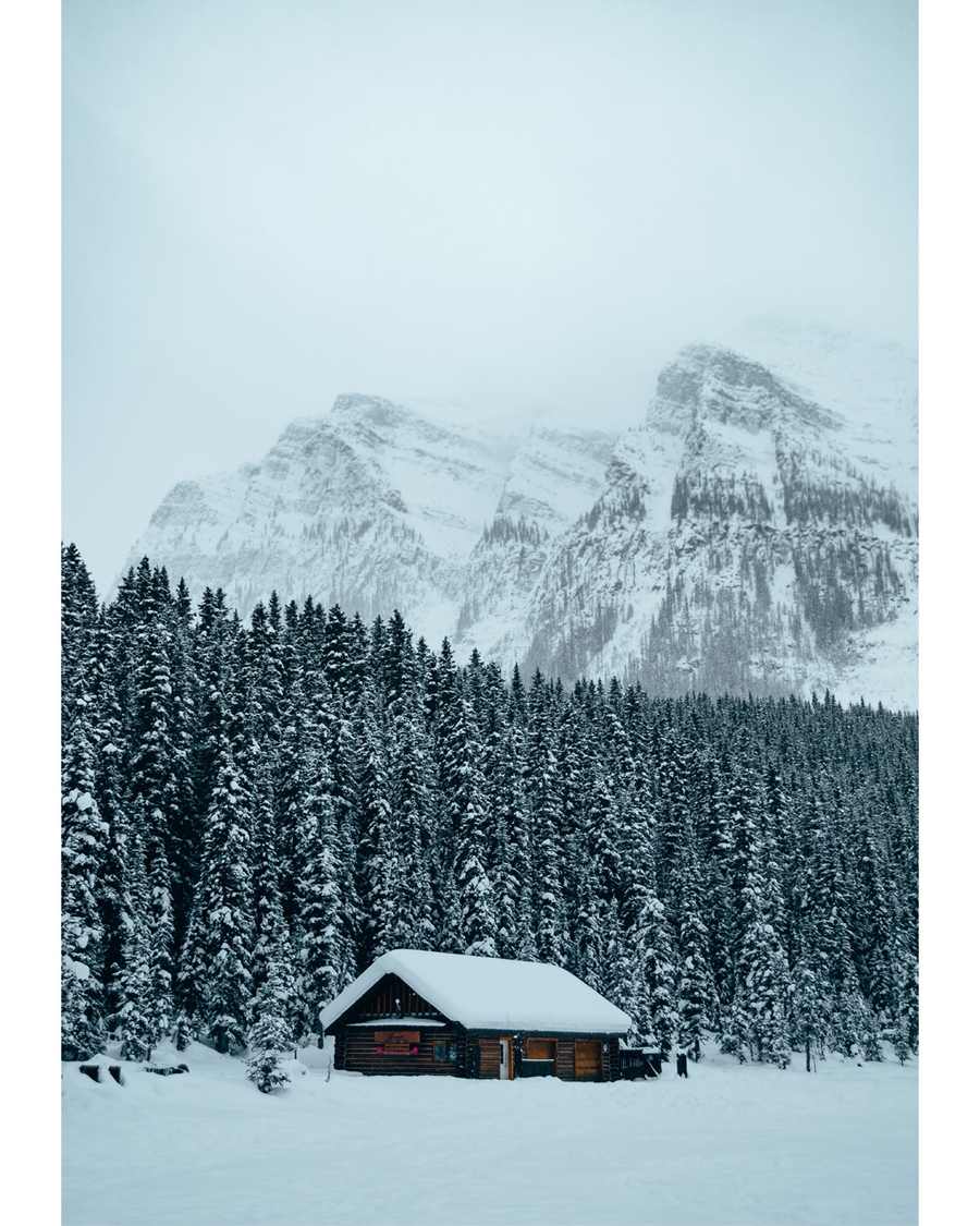 Snow-covered cabin surrounded by pine trees and mountains