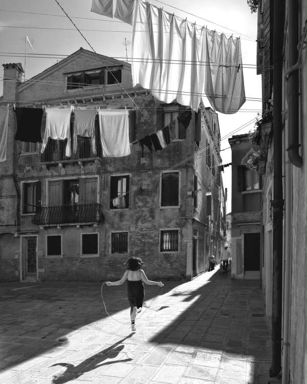 Black and white street photo of a girl skipping rope in a sunlit square under laundry lines