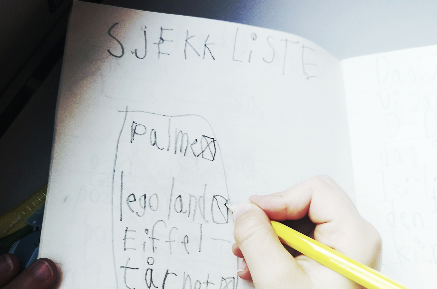 En kladdebok der et barns hånd skriver en sjekkliste over ting som skal oppleves på turen. Sjekklisten inneholder palmer, lego-land og eiffel-tårnet.