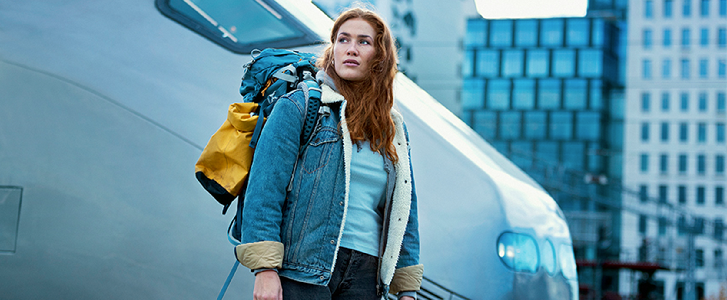 En ung kvinne står ved siden av fronten på et tog. I bakgrunnen er det høye byggninger.