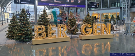 Bilde av juledekorasjon på Bergen stasjon. Det er juletrær med lys, og store opplyste bokstaver som staver Bergen. I bakgrunnen sees et tog i stasjonshallen