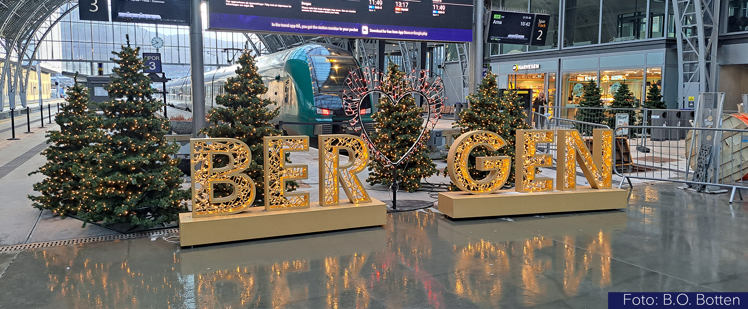 Bilde av juledekorasjon på Bergen stasjon. Det er juletrær med lys, og store opplyste bokstaver som staver Bergen. I bakgrunnen sees et tog i stasjonshallen