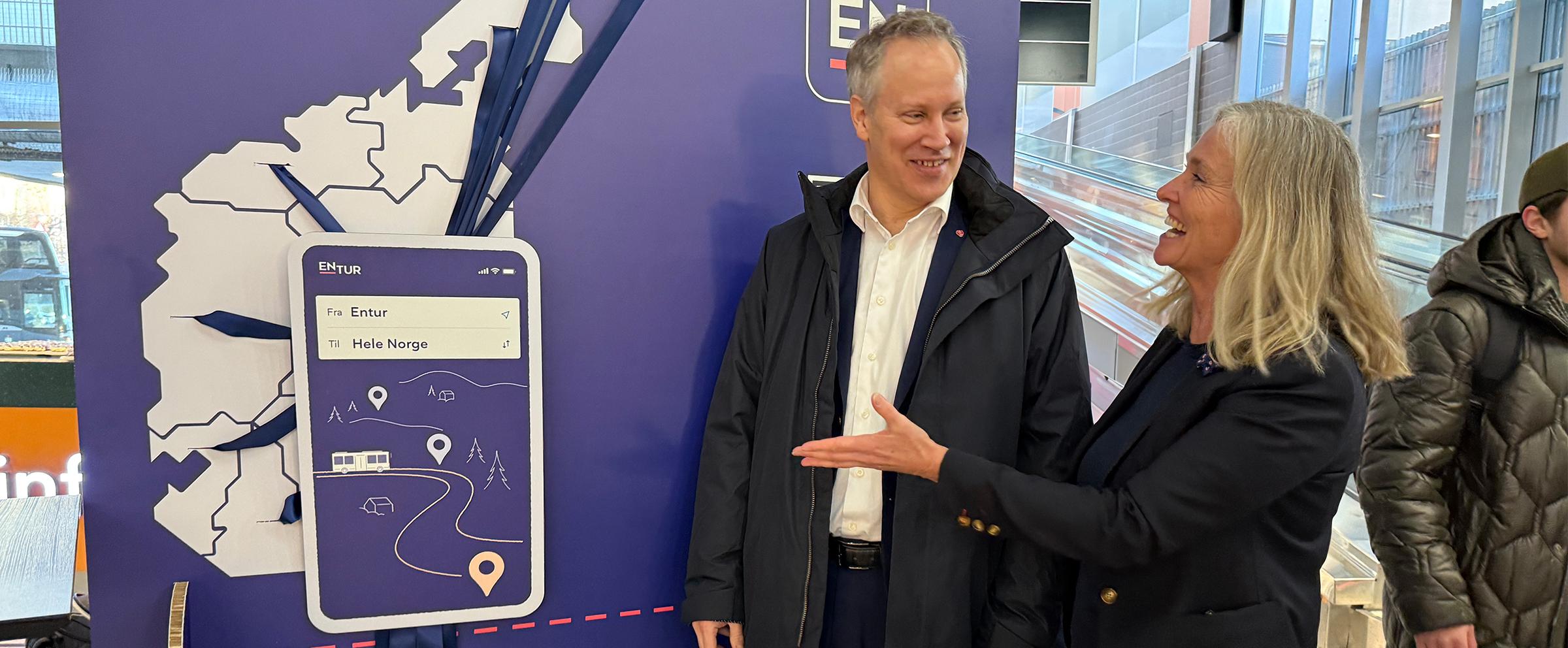 Bilde av adm.dir Borge og samferdsminister Nygård som står foran en plakat av et norgeskart. De smiler til hverandre og Borge peker på plakaten. På norgeskartet ser man silketråder som strekker seg fra hvert fylke inn mot en stor papp-mobil. "Skjermbildet" på mobilen er Entur-appen, der det står "Fra Entur, Til Hele norge"