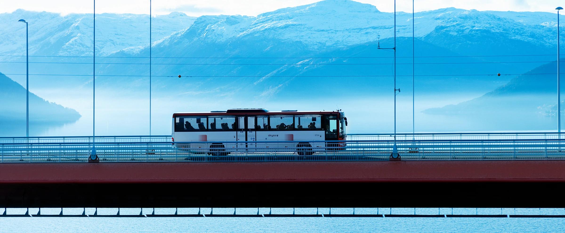 En hvit buss krysser en hengebro over en bred fjord. I bakgrunnen synes et blånende fjellmassiv Normalperspektiv gjør at broa danner en linje gjennom bildet. Foto