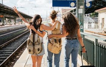 Bilde av tre venninner som holder rundt hverandre og står ved siden av togskinner med sekker og bagasje fullpakket.