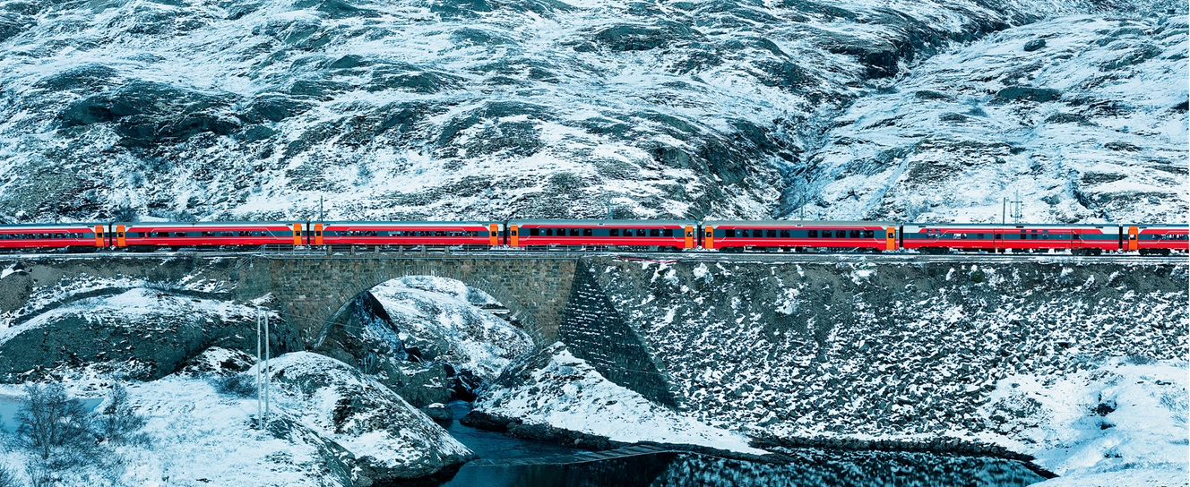 Et rødt tog krysser en gammel steinbro over en elvemunning. Elven renner ut i et stort vann som utgjør bildets nederste tredjedel. I øverste tredjedel synes en snødekt fjellside. Bildet har normalperspektiv, og toget danner en linje gjennom hele komposisjonen.