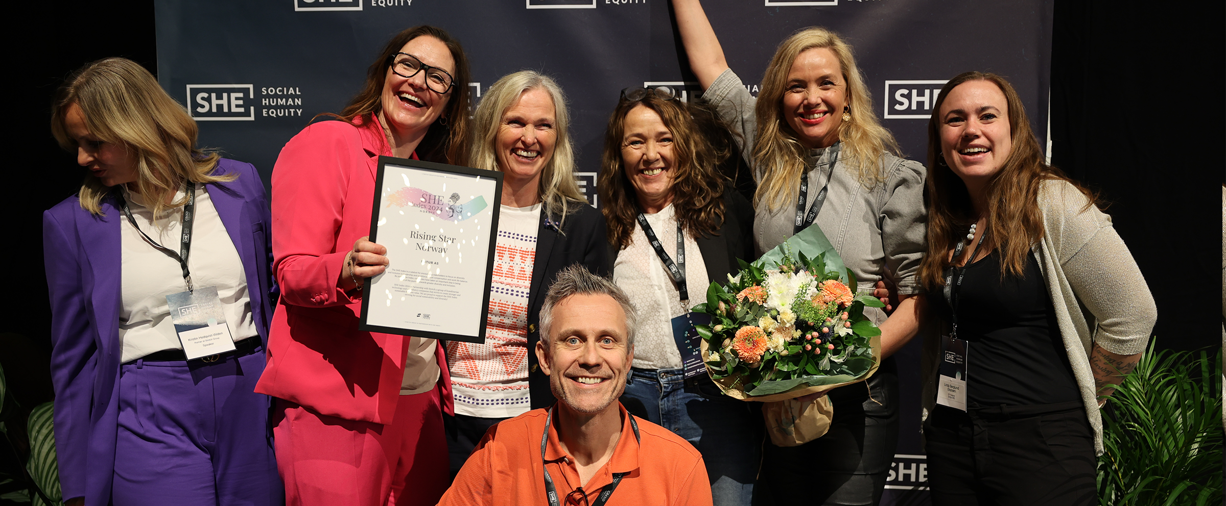 Bilde av enturs representanter ved She Conference som står forran en fotovegg og jubler. Diplom og blomsterbukett.