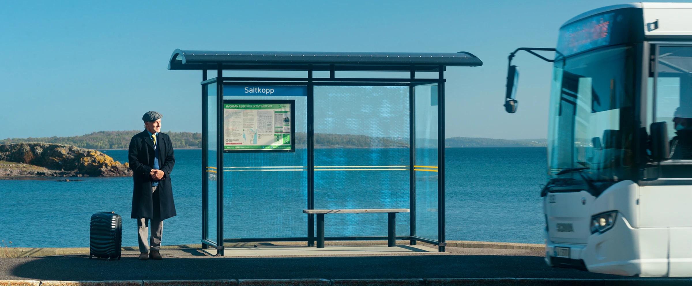 En mann står ved stoppestedet Saltkopp. Han har koffert, og bussen er akkurat på vei til å stoppe.