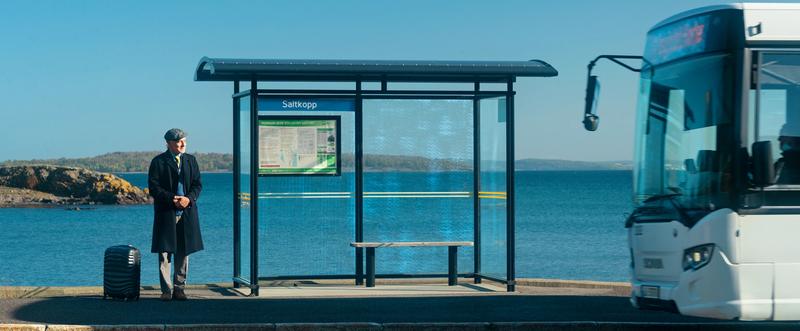 En mann står ved stoppestedet Saltkopp. Han har koffert, og bussen er akkurat på vei til å stoppe.