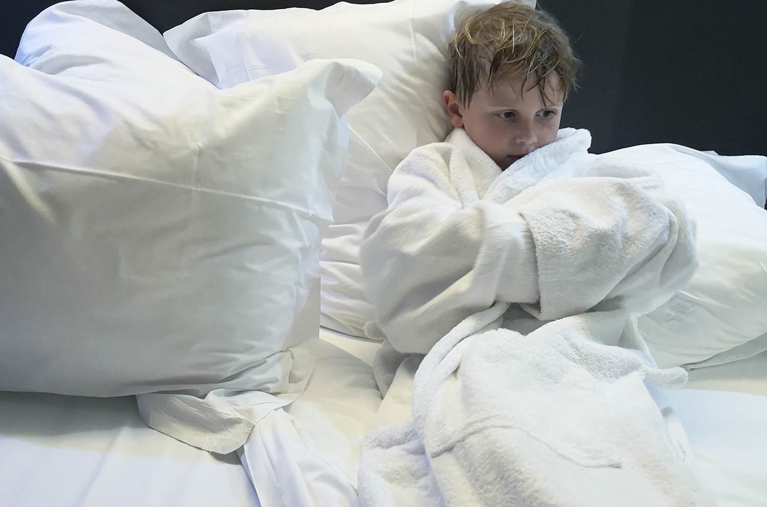 Sønnen sitter i en seng med badekåpe og luksus-sengetøy