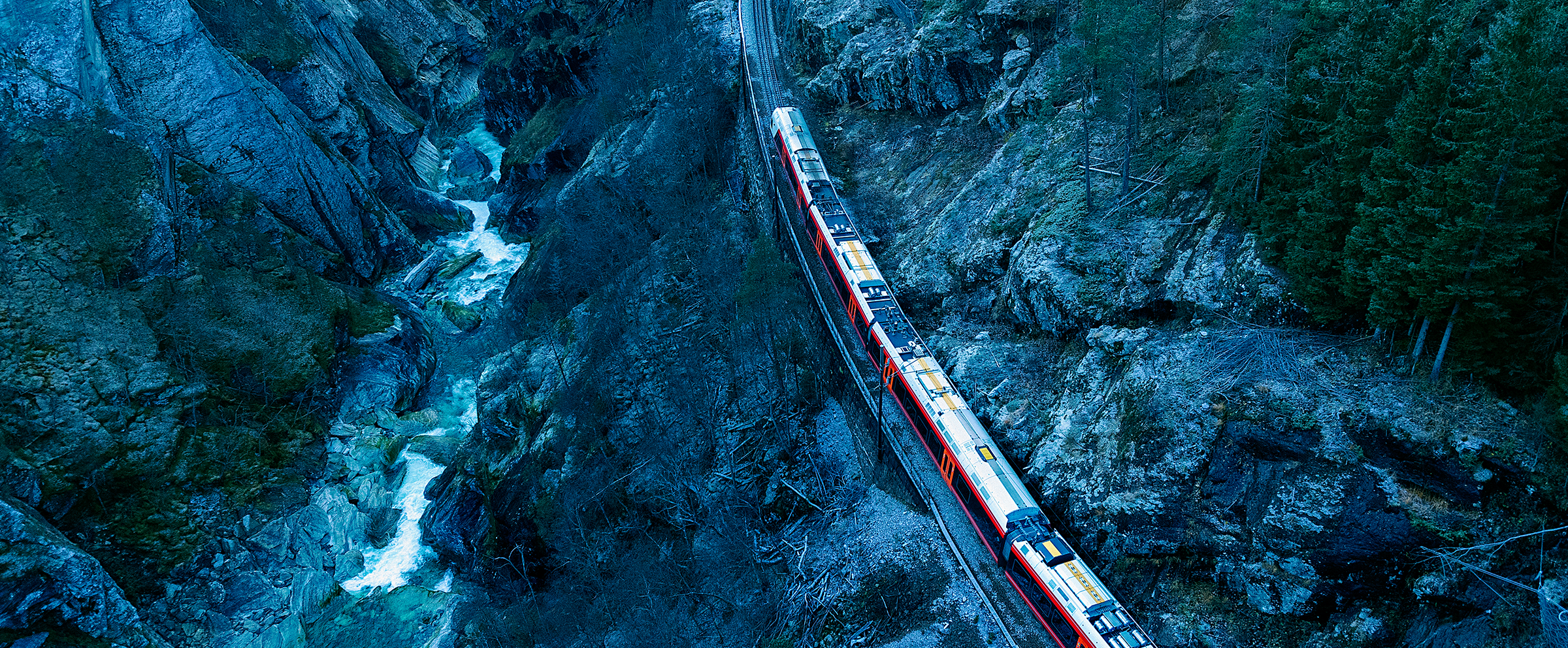 Foto: Et tog kjører snart inn i en tunnel i et svært bratt berg. Til venstre i bildet renner en elv i et juv. Jernbanen ligger parallelt med elveleiet, men lenger opp i skrenten. Toget kommer fra nederste høyre kant av bildet og kjører mot tunnelen som ligger lengt opp og i senter av bildet.